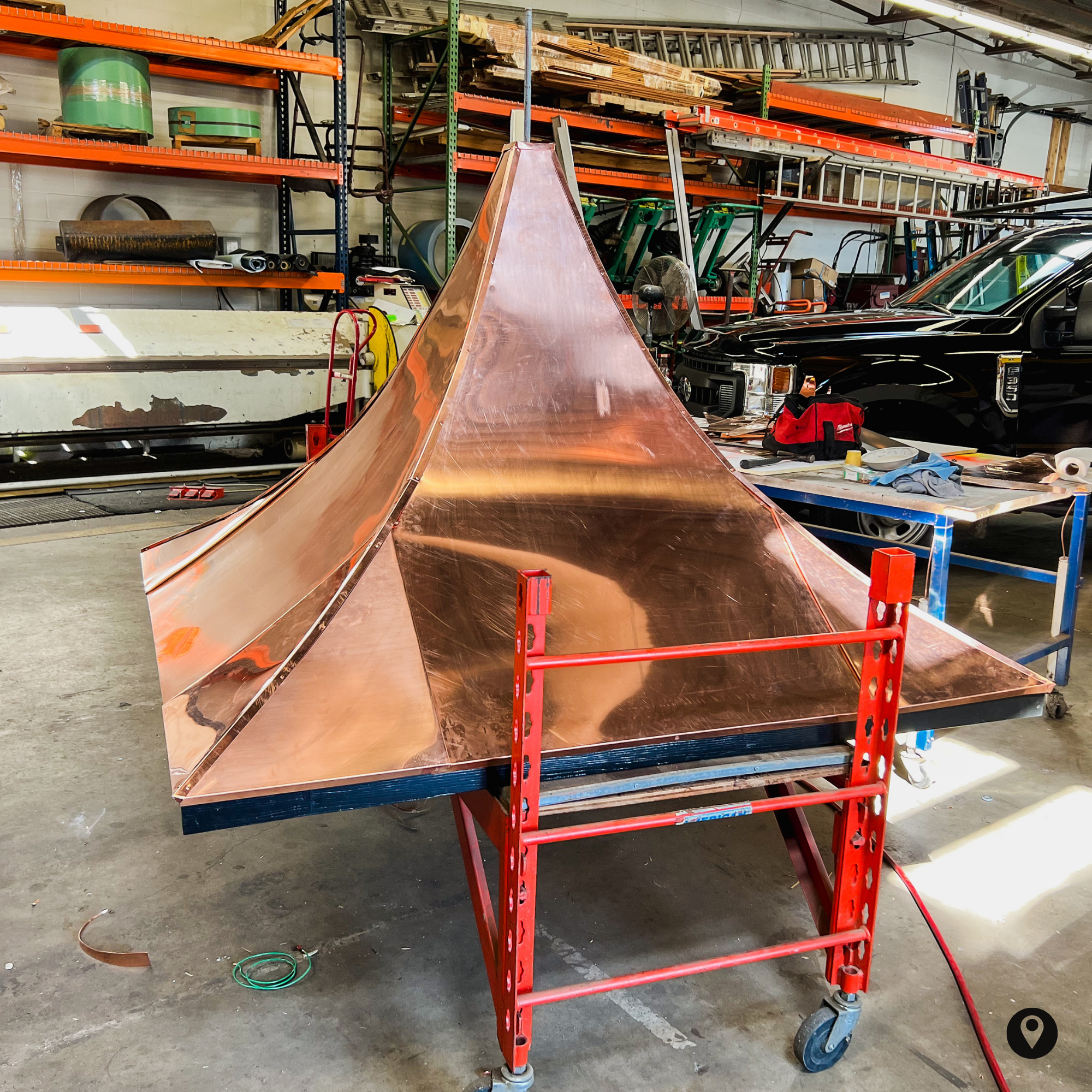 A workshop or warehouse with orange shelving holding wood, metal, and ladders. There is a copper-colored shaped object, possibly a piece of metalwork, supported by a red dolly with wheels. Tools and equipment are scattered around on the ground and shelves.