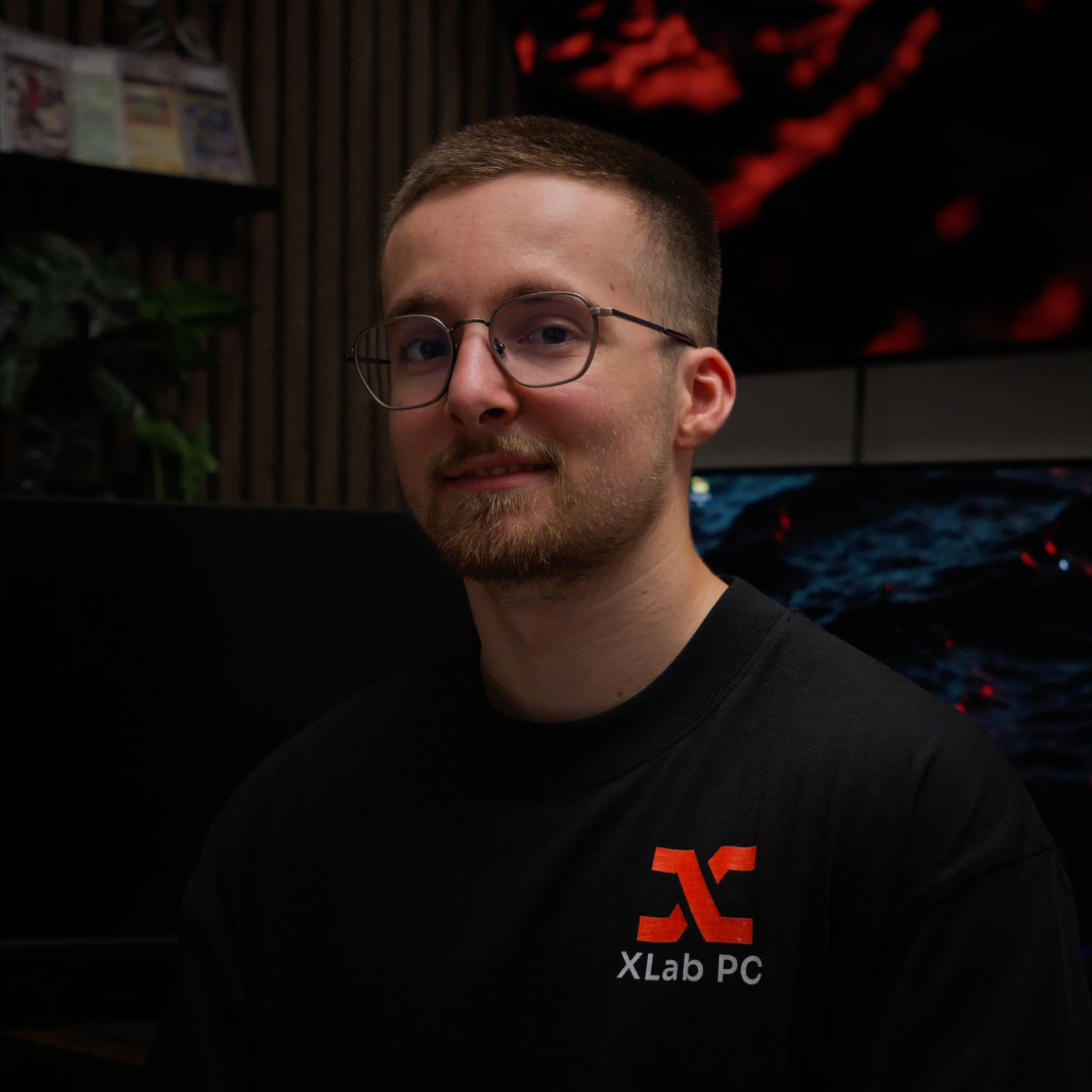 Jeune homme portant des lunettes, T-shirt noir avec lettres orange, se trouvant dans un environnement intérieur avec décor style bois et écran d'ordinateur en arrière-plan.