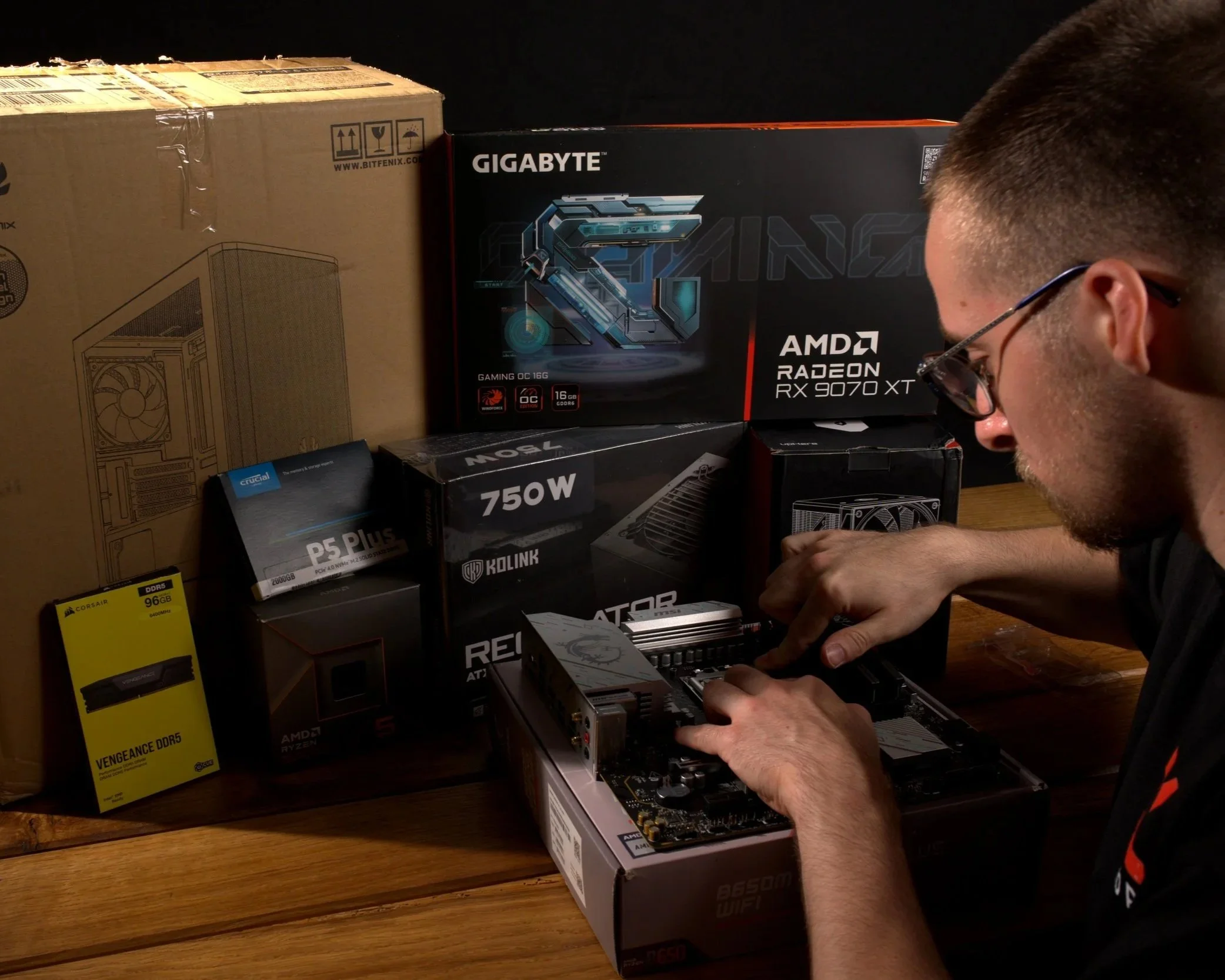 Une personne assemble un composant informatique dans une pièce avec des boîtes et cartons de matériel informatique autour, notamment une carte graphique AMD Radeon RX 9070 XT, une alimentation de 750W, et des modules de mémoire RAM DDR5.