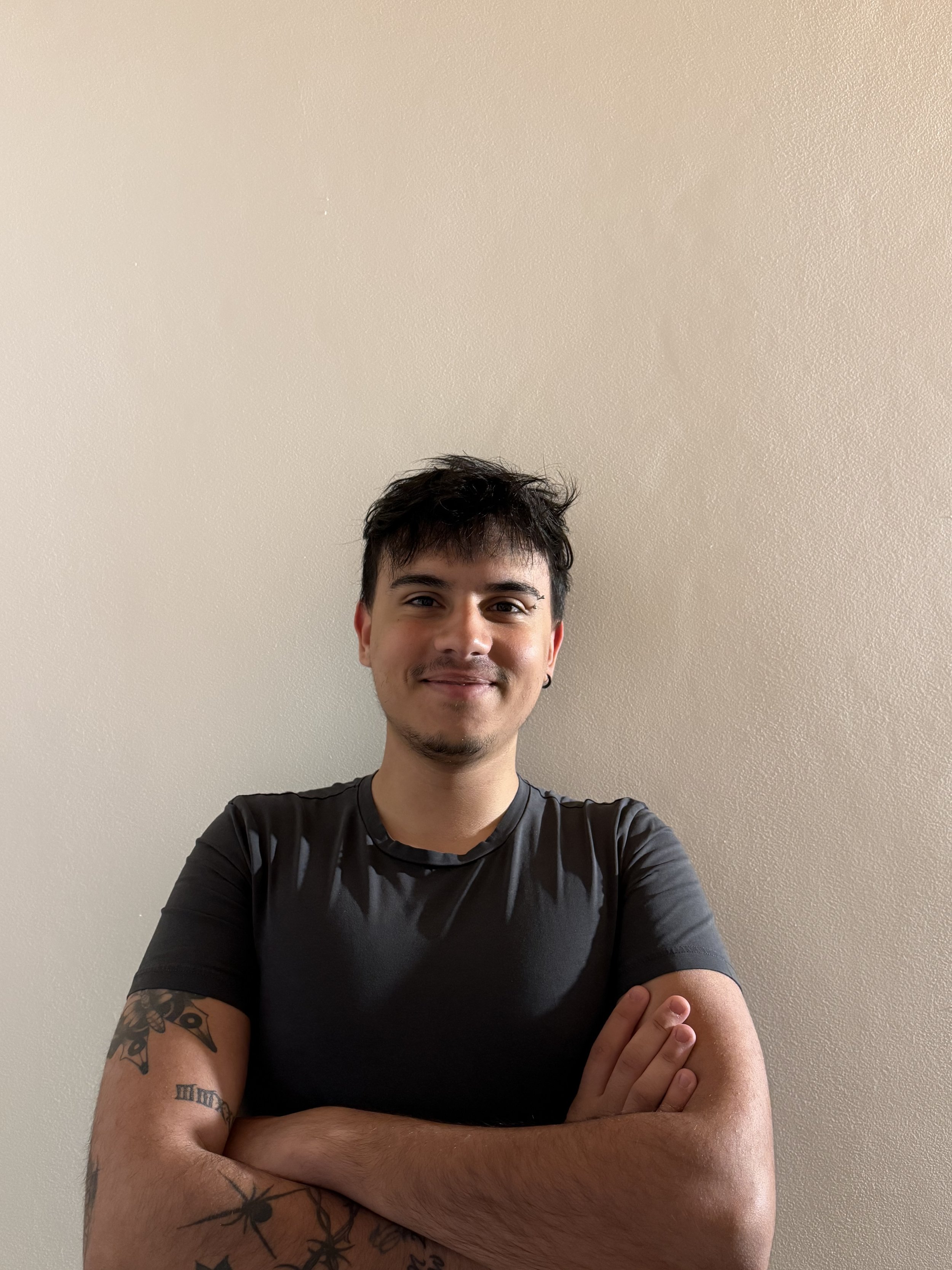 A young man with his arms crossed, smiling and wearing tattoos, against a beige wall.