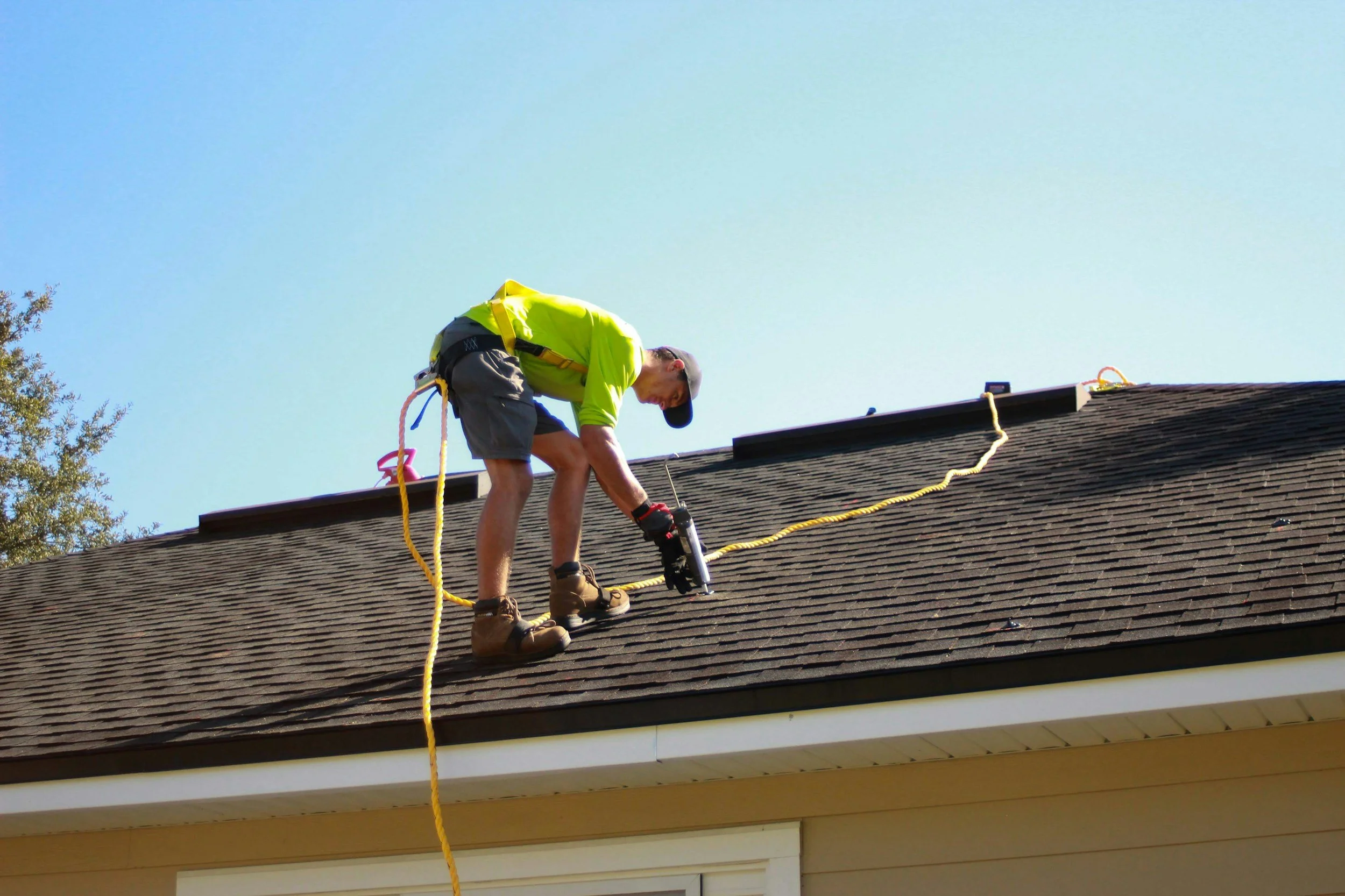 A man in a gray hat and yellow safety vest is fixing a rooftop while using climbing gear and a yellow rope.