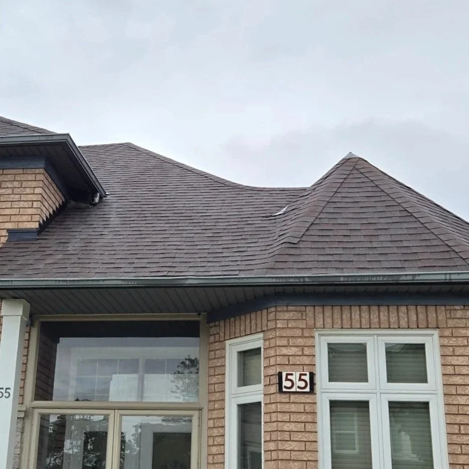Close-up of a brick house's roof with a peak, brick walls, and a window with blinds visible.