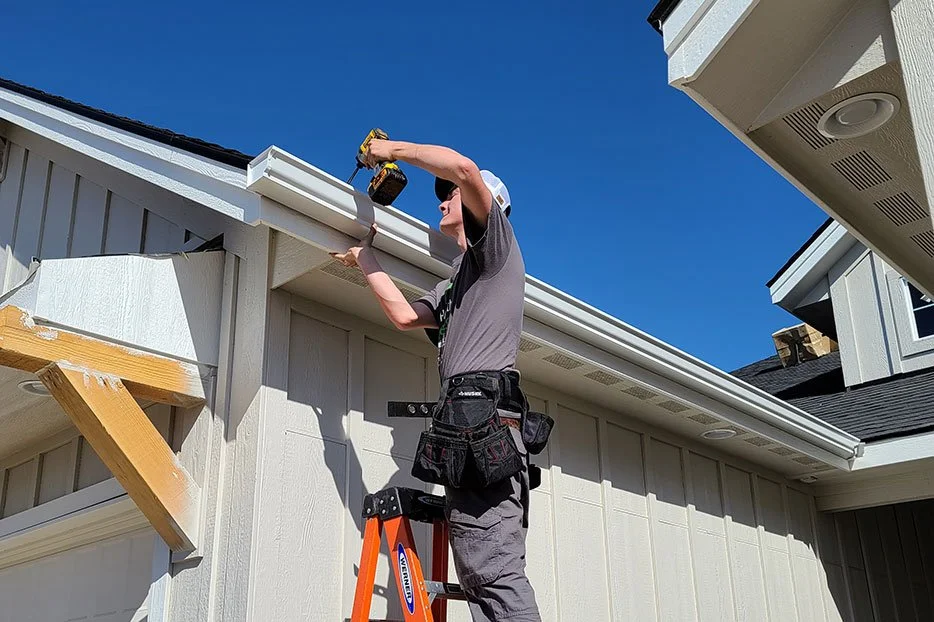 A man standing on an orange ladder installing or repairing a gutter on a white house, with a clear blue sky in the background.