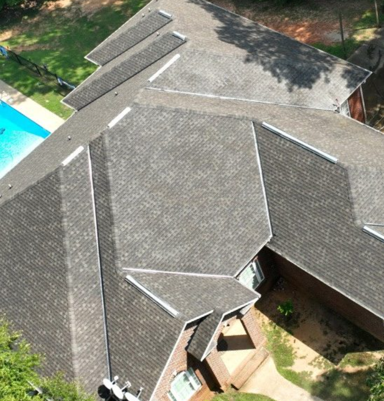 Aerial view of a house with a complex, multi-section shingled roof, neighboring yard, and a swimming pool in the backyard.