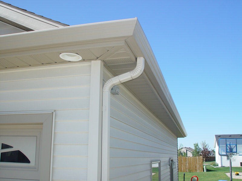 Outdoor view of a house's white vinyl siding, corner gutter system, and a small outdoor light fixture under the roof overhang, with a neighboring house and a fenced backyard in the background.