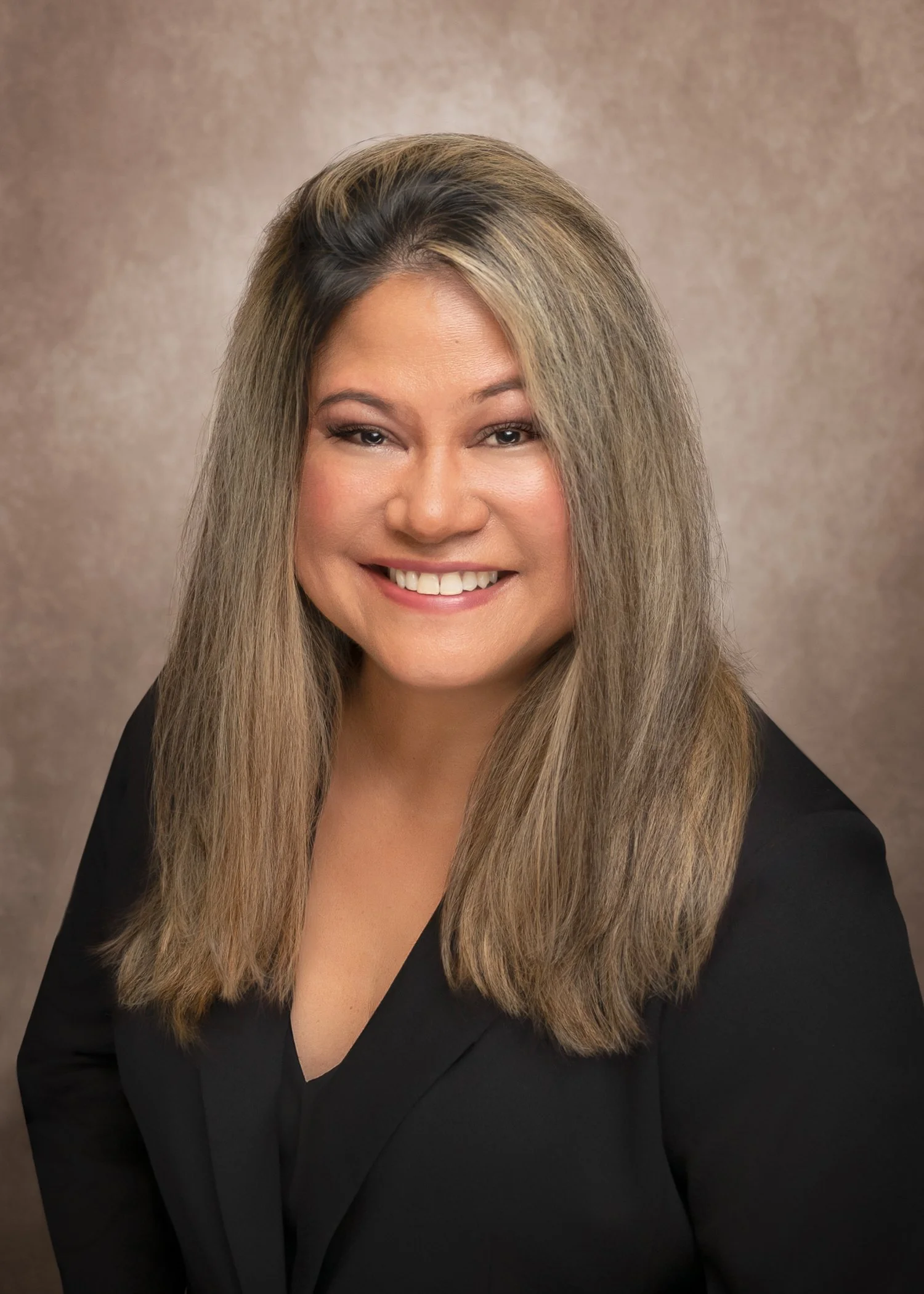 A woman with long, styled hair wearing a black blazer, smiling, against a neutral background.