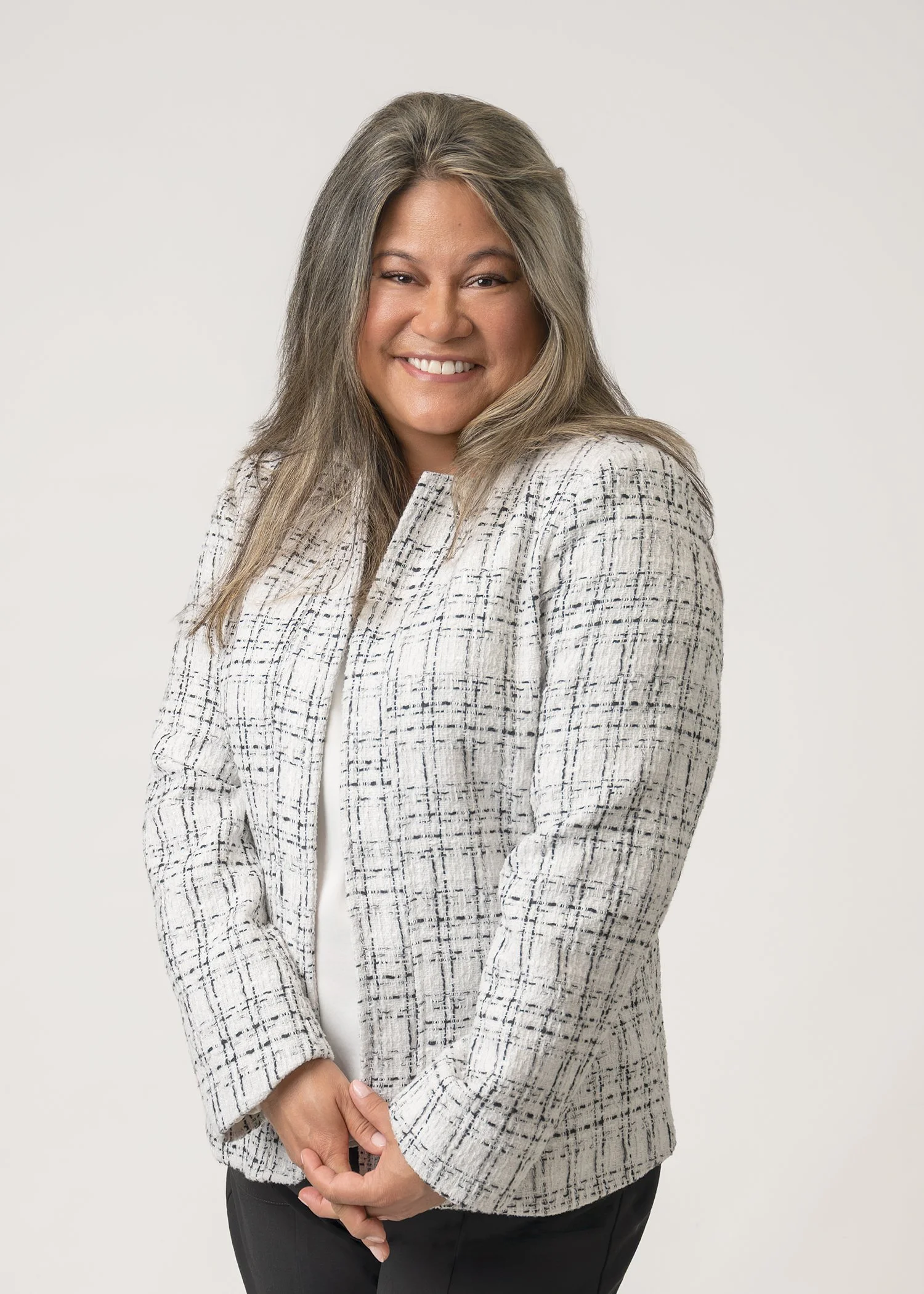 A woman smiling, wearing a white blazer with black checkered patterns and black pants, against a plain white background.