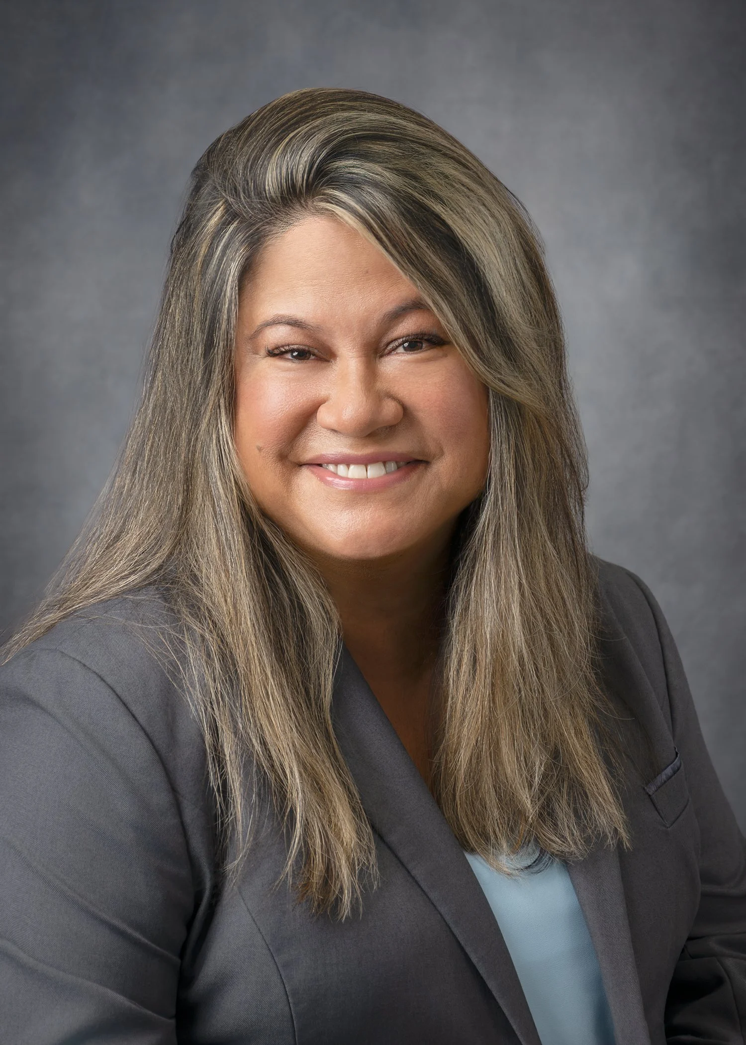 Professional woman with long hair smiling, wearing a dark blazer and light blue shirt against a gray background.