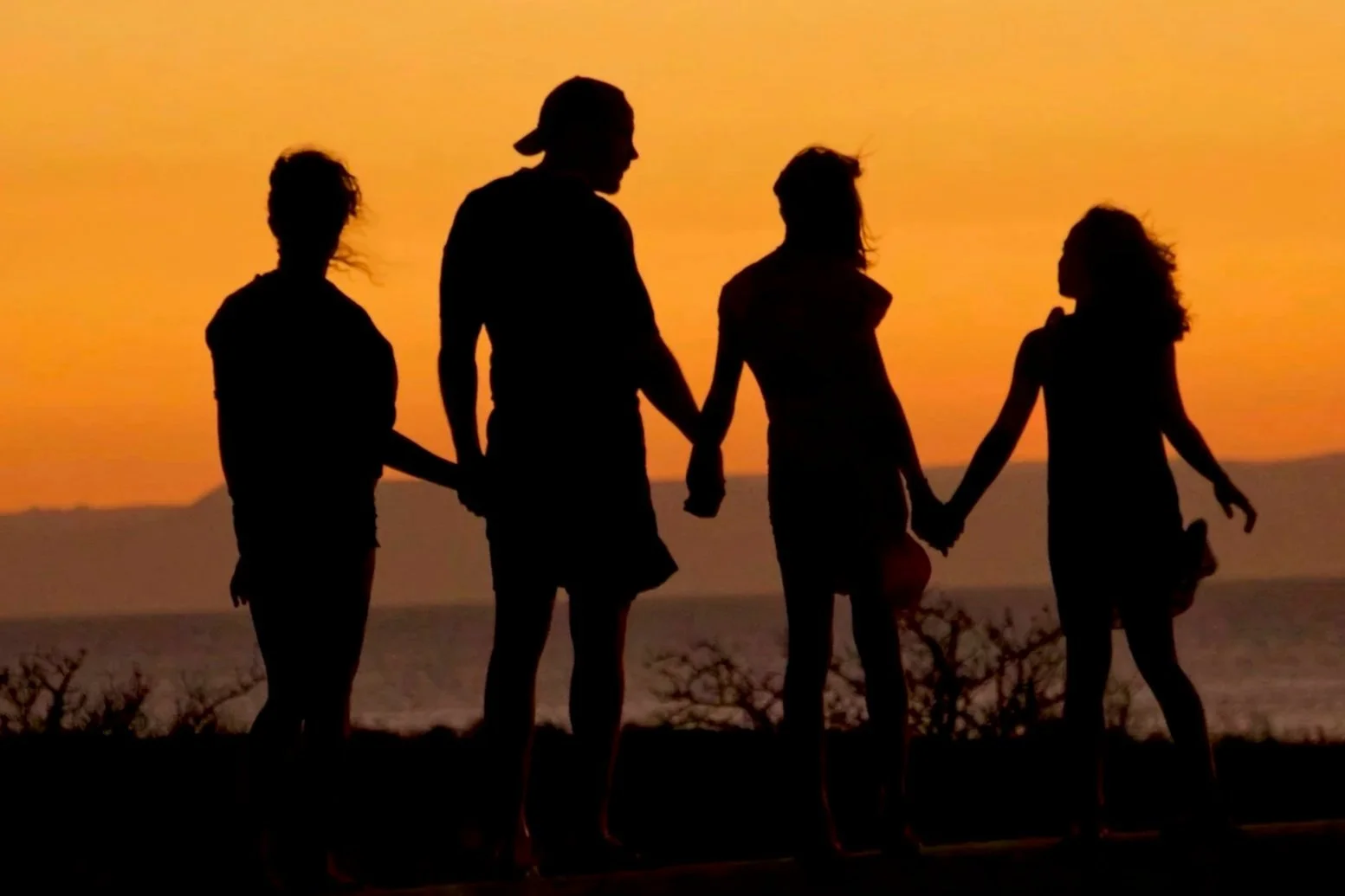 Silhouettes of four people holding hands at sunset near a body of water.