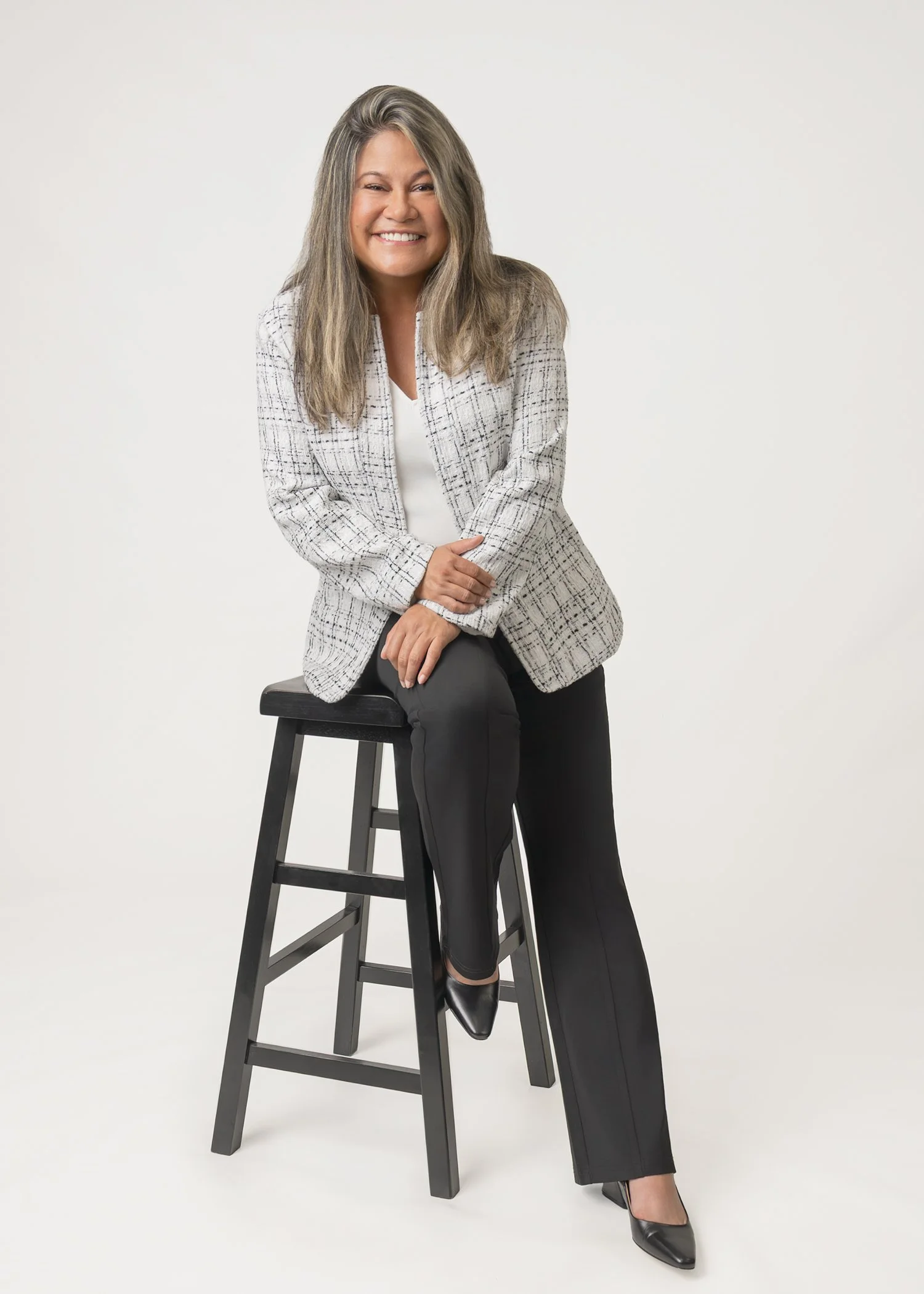 A woman with long, wavy blonde hair, wearing a light-colored, plaid blazer, white top, black wide-leg pants, and black heels, sitting on a tall black stool with one leg crossed over the other, smiling at the camera against a plain white background.