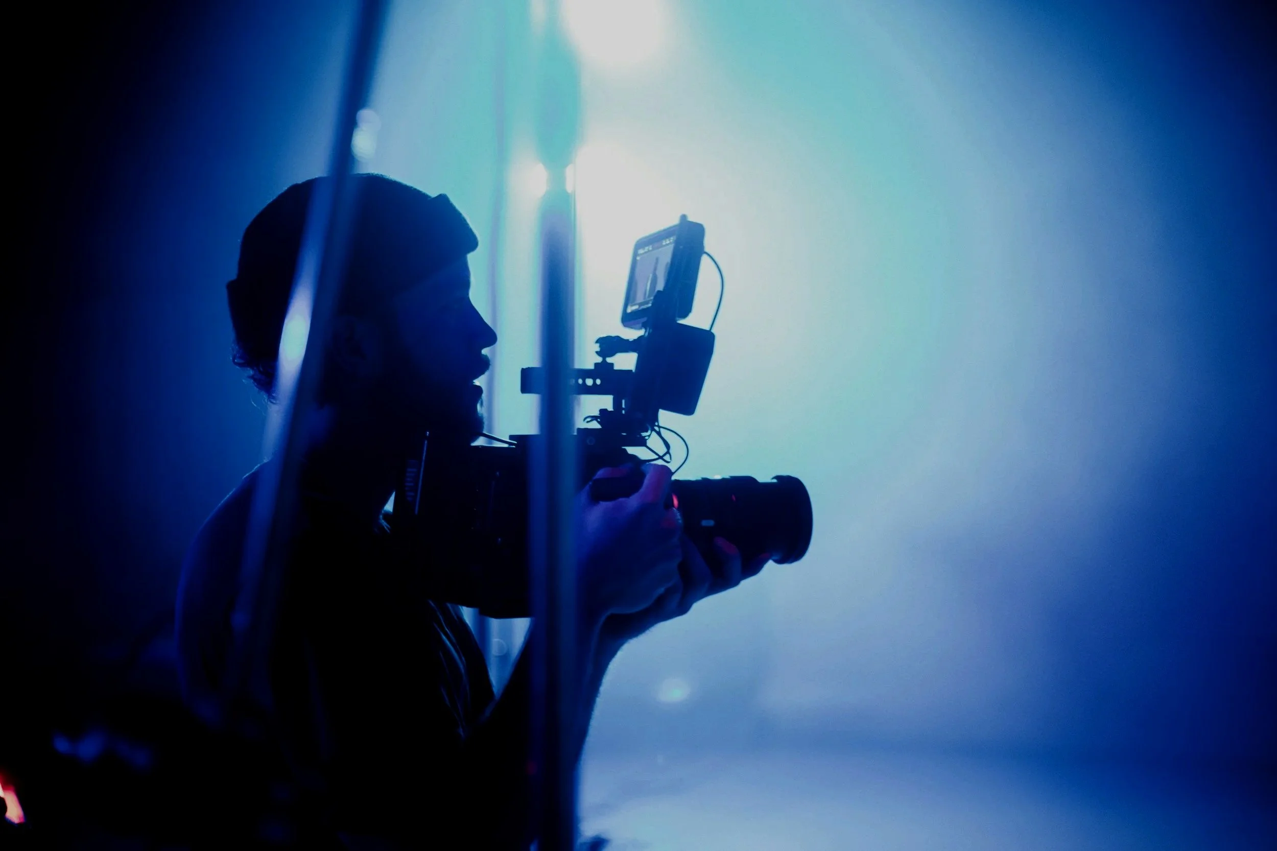 Silhouette of a person operating a camera or filming device against a bright blue and white backlight.