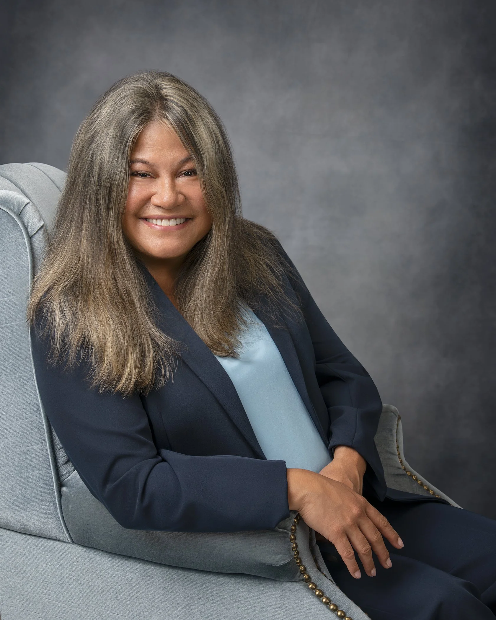 A smiling middle-aged woman with long, light brown hair with gray streaks, seated on a gray upholstered armchair, wearing a navy blazer and light blue blouse, posed against a gray backdrop.