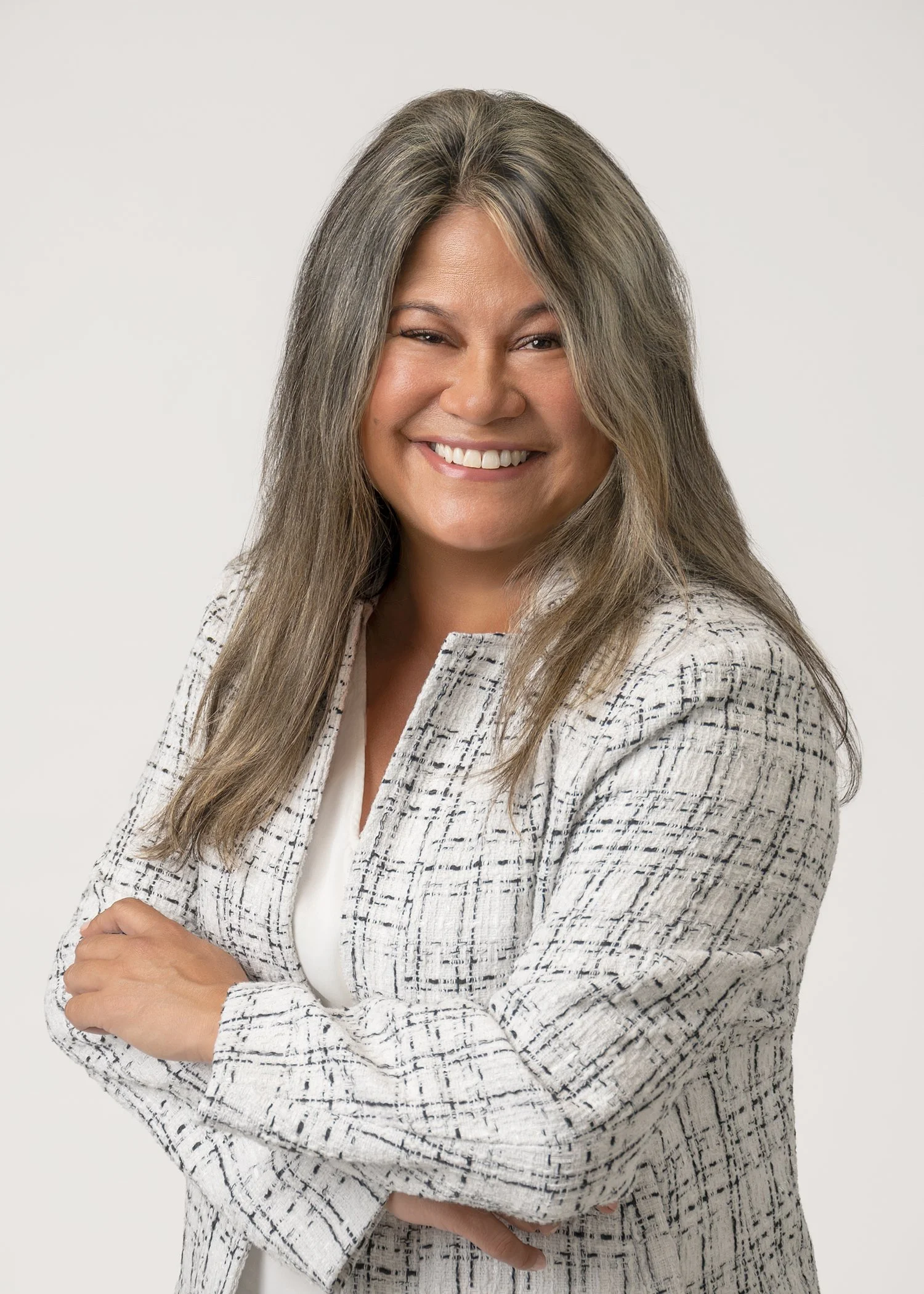 A woman with long, wavy gray hair smiling and winking, wearing a white outfit and a white and black checkered blazer, with arms crossed in front of a plain white background.