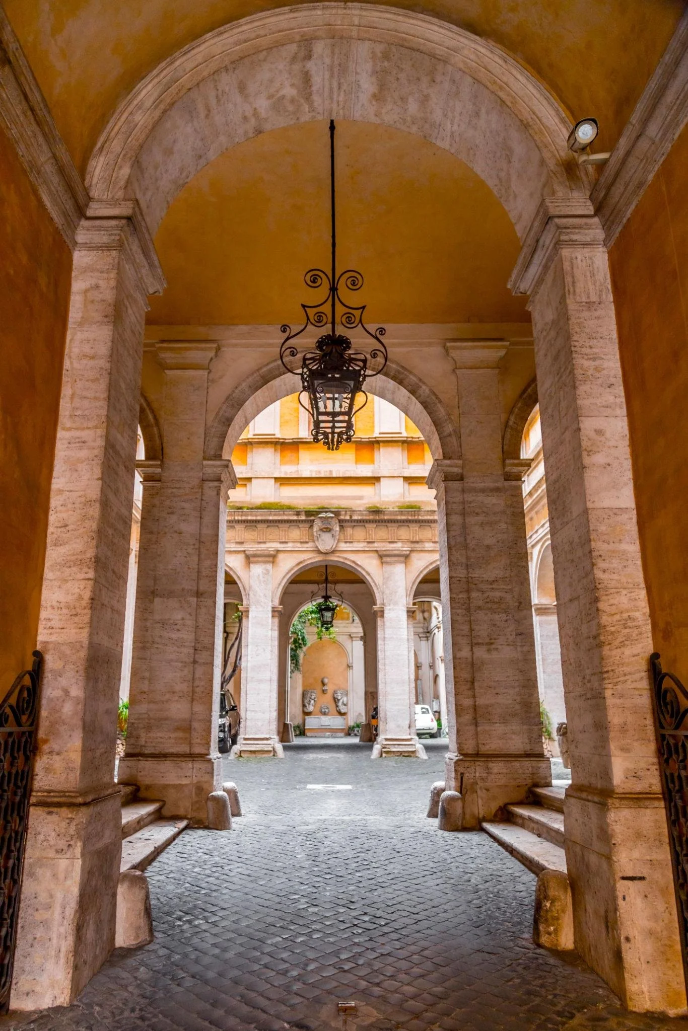 A view through an old Italian arched passageway in a historic building, showing stone columns, a decorative wrought iron chandelier, and a courtyard with parked cars.