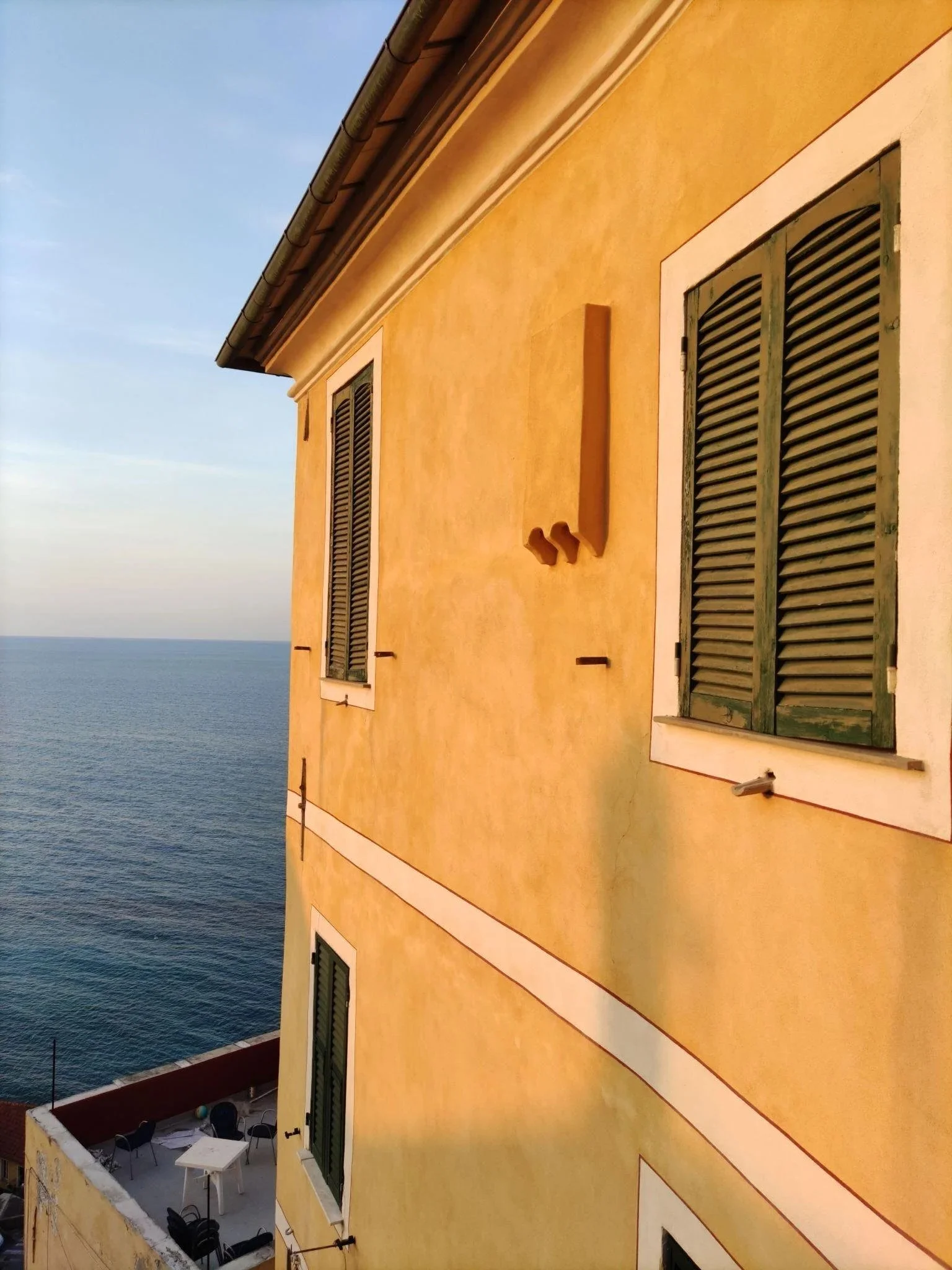 Yellow building with green shutters on windows, overlooking the ocean under a clear blue sky. Boccadasse, Nervi, Chiavari or similar area