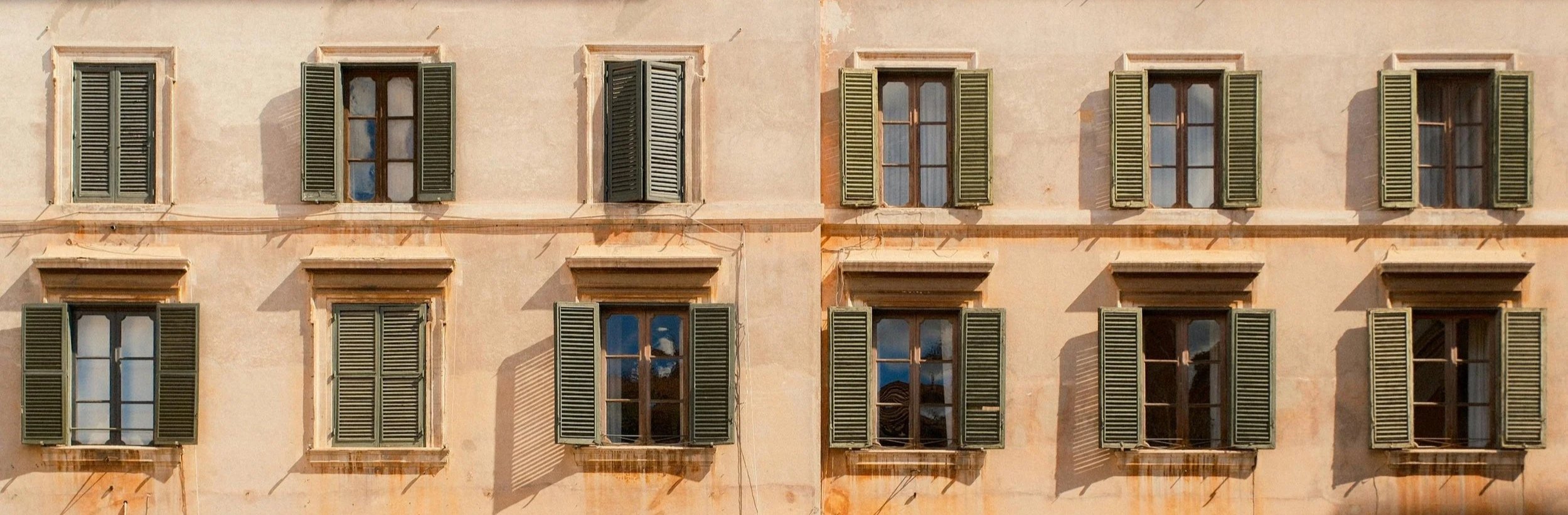 A building facade in Liguria, Genova with nine windows, three per row, with green shutters, some open and some closed, and a light peach-colored in aged plaster wall with shadows cast by the shutters.