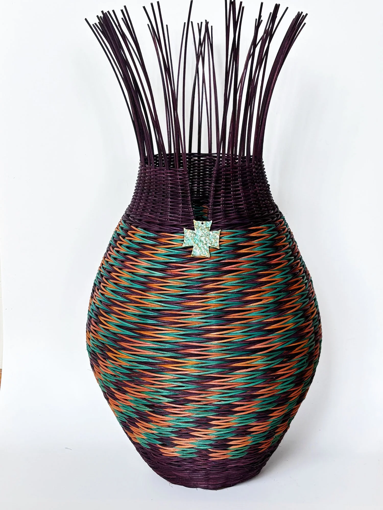 A colorful woven basket with a cross-shaped ornament attached to the front, featuring a blend of purple, orange, green, and black strands, against a plain white background.