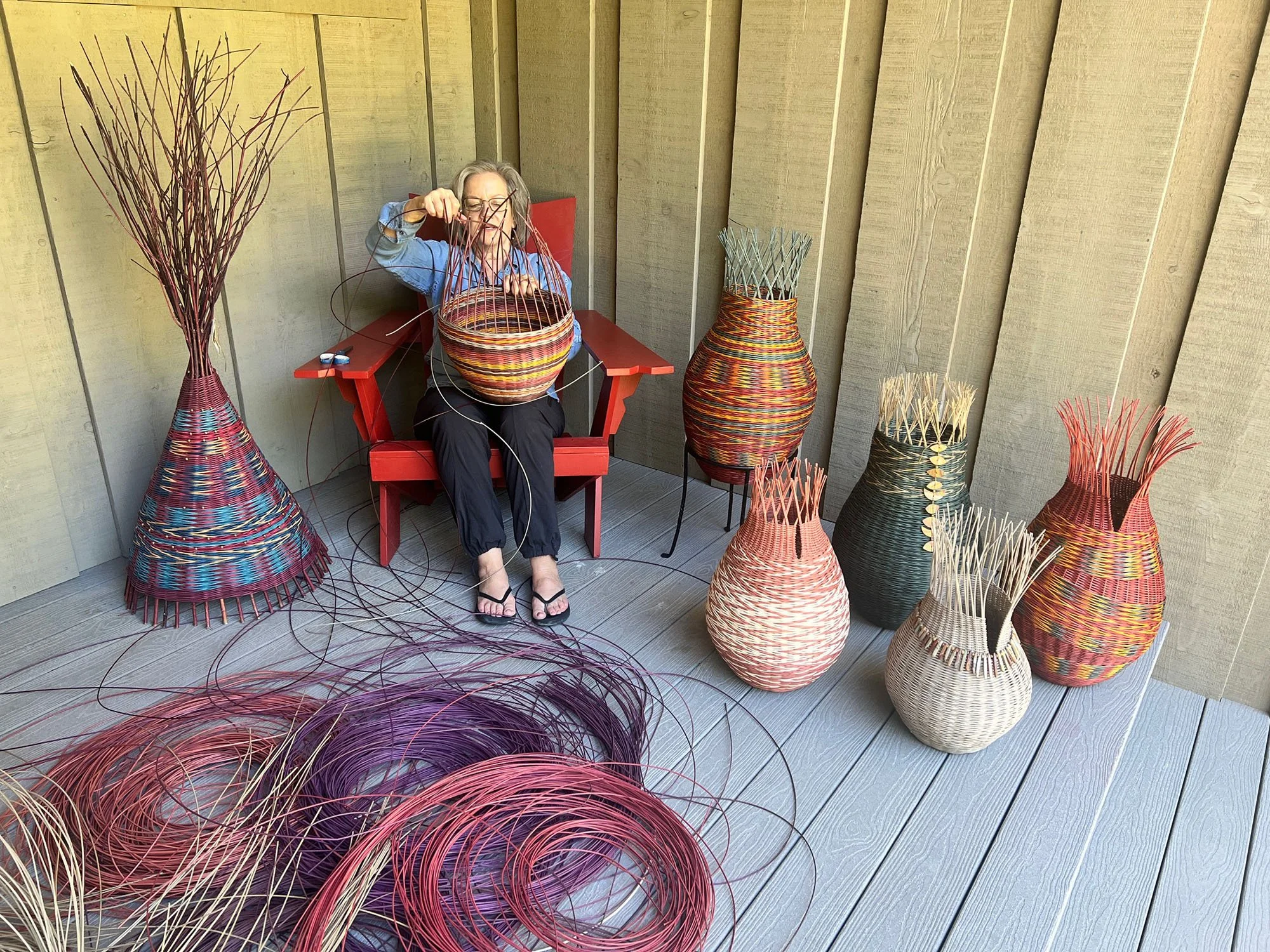 The artist sitting on a red chair weaving a basket surrounded by finished woven baskets and large decorative woven vases on a wooden deck with a beige wooden wall in the background.
