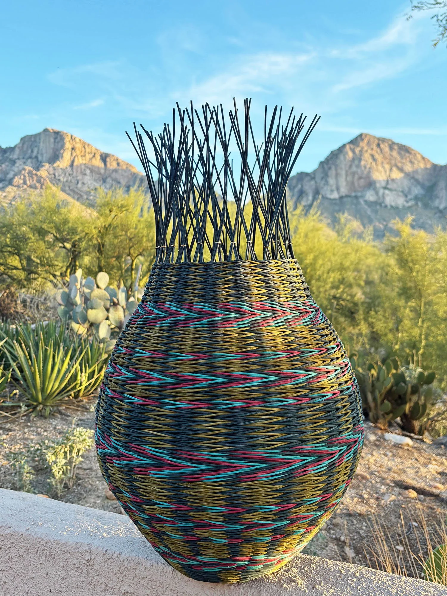 Blue and yellow green woven pot with prominent multi-colored arrows. Finished with reed spokes resembling fringe.