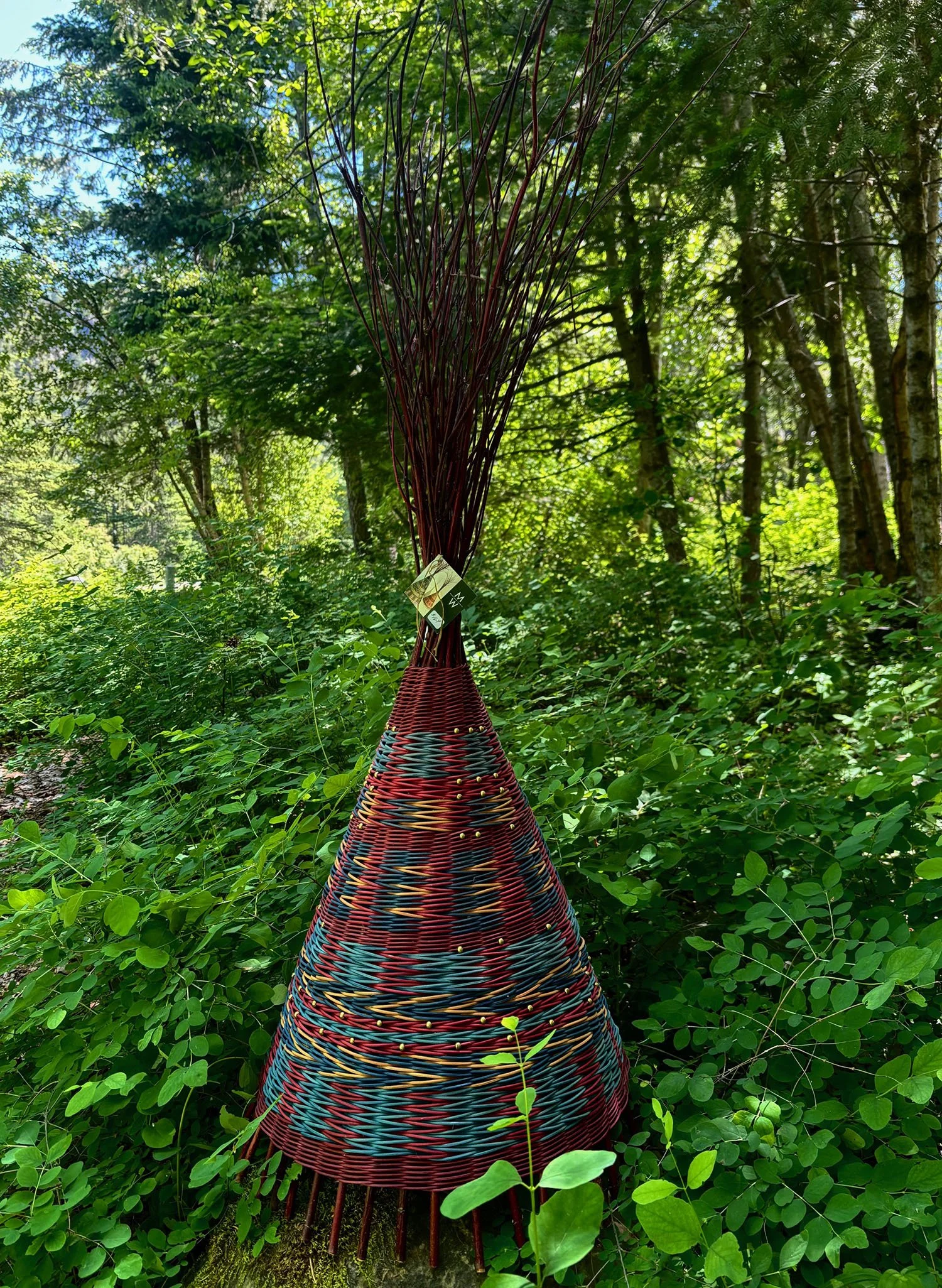 A colorful woven sculpture in an outdoor wooded area with tall trees and green foliage, featuring a conical shape resembling a "tipi".