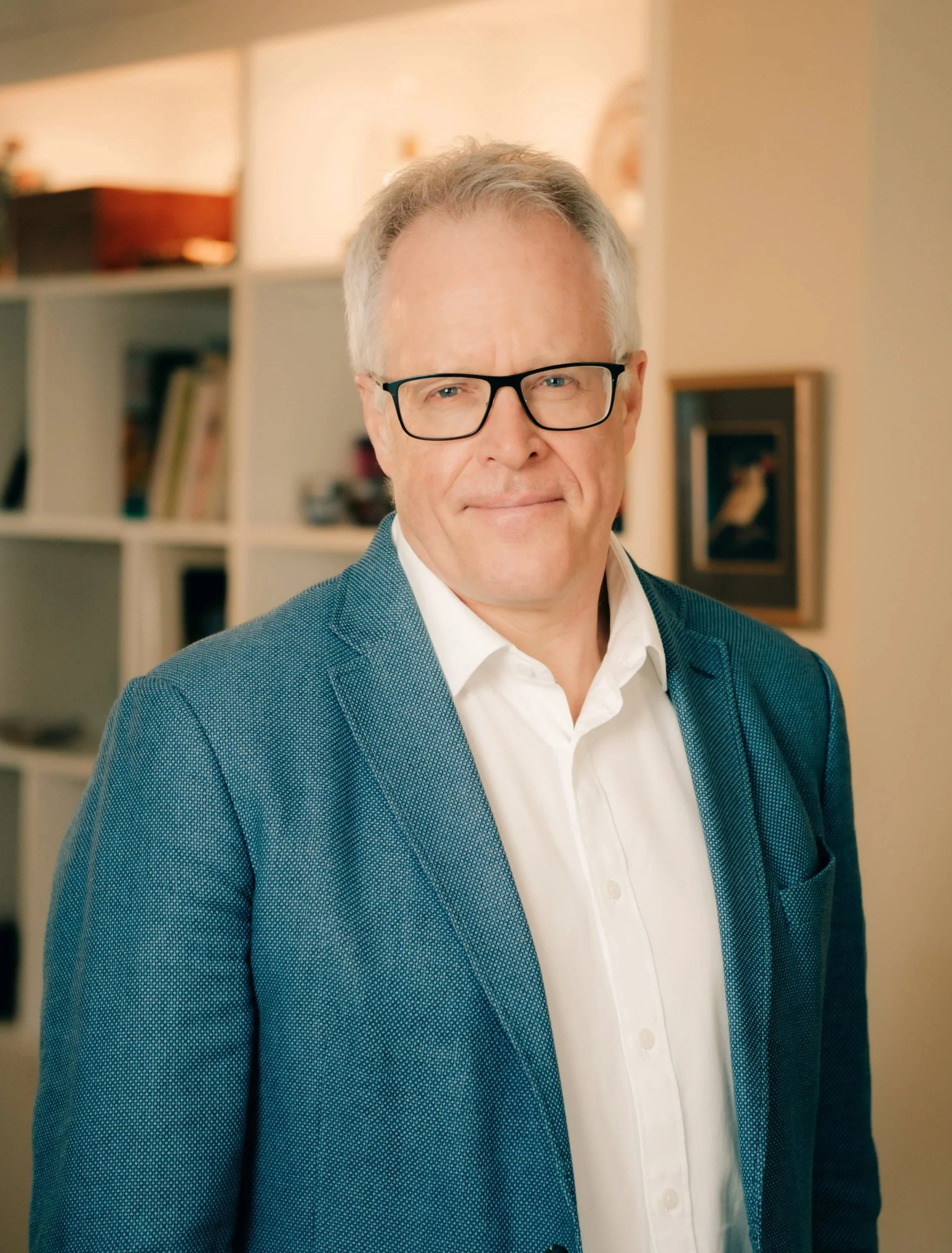 A middle-aged man with gray hair wearing glasses, a blue blazer, and a white shirt, standing indoors in front of a blind bookshelf with blurred framed art on the wall.