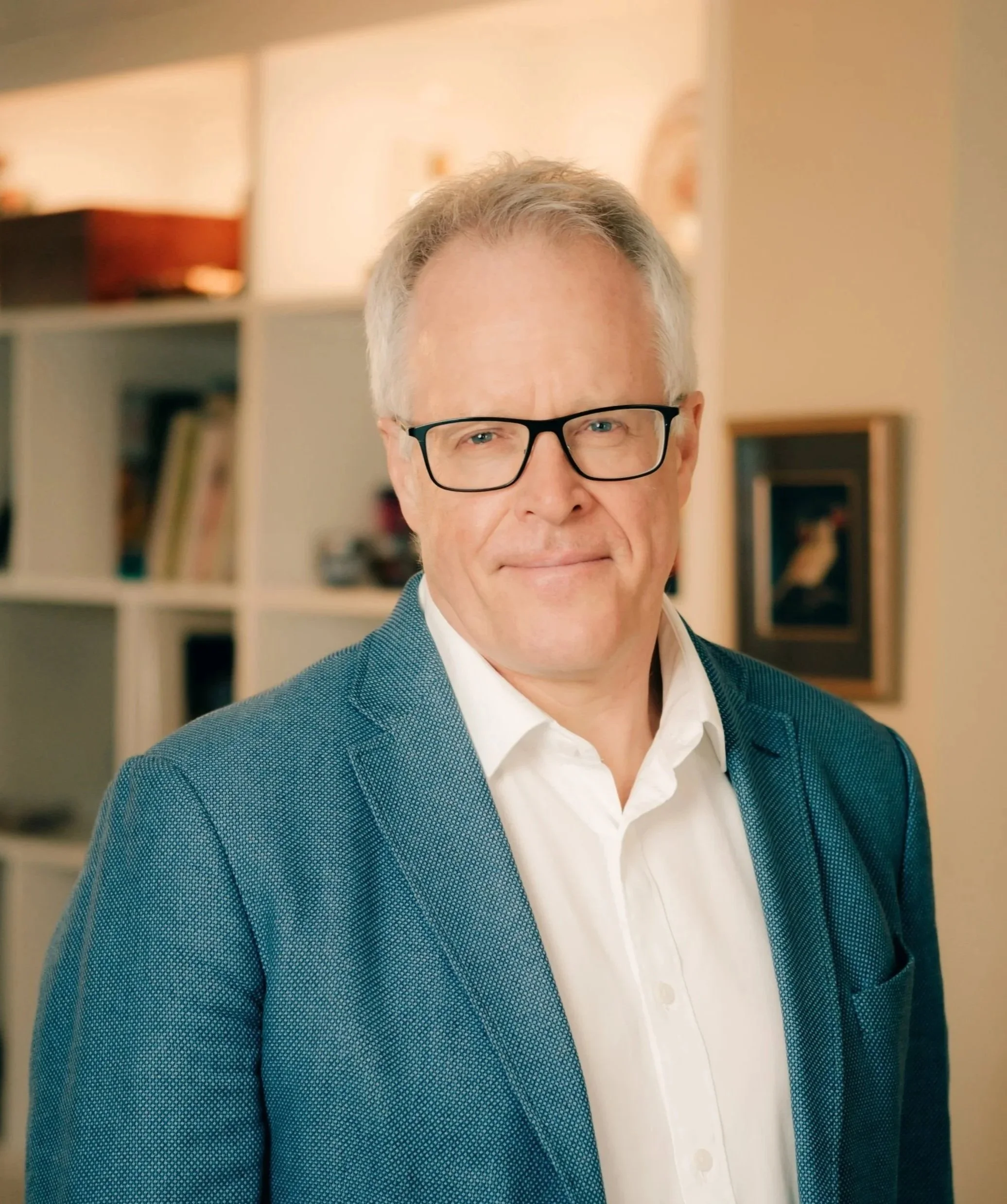 A  man with grey hair and glasses, wearing a blue blazer and white shirt, standing indoors with a blurred bookshelf and artwork in the background.