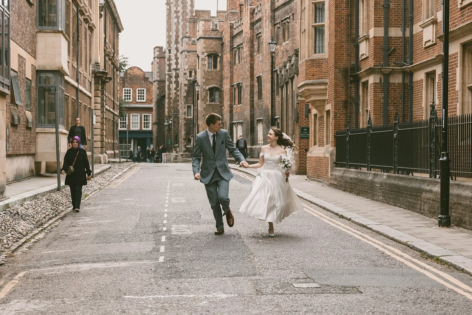 A newlywed couple holding hands and running in the middle of a street with old brick buildings on either side. The bride is wearing a white wedding dress, and the groom is in a suit.