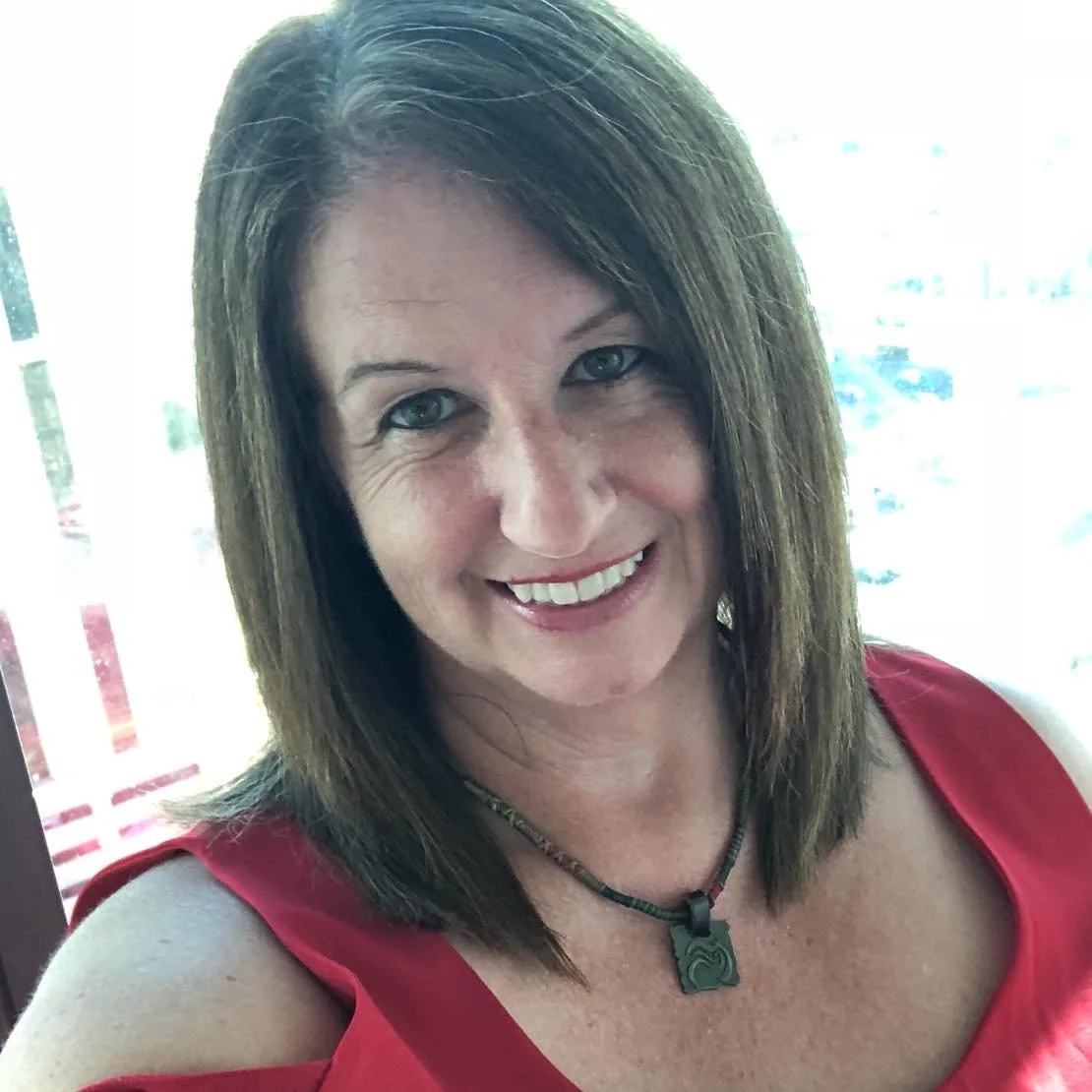 A woman with shoulder-length brown hair, wearing a red top and a necklace, smiling at the camera, standing indoors near a window with a blurred view of the outdoors.