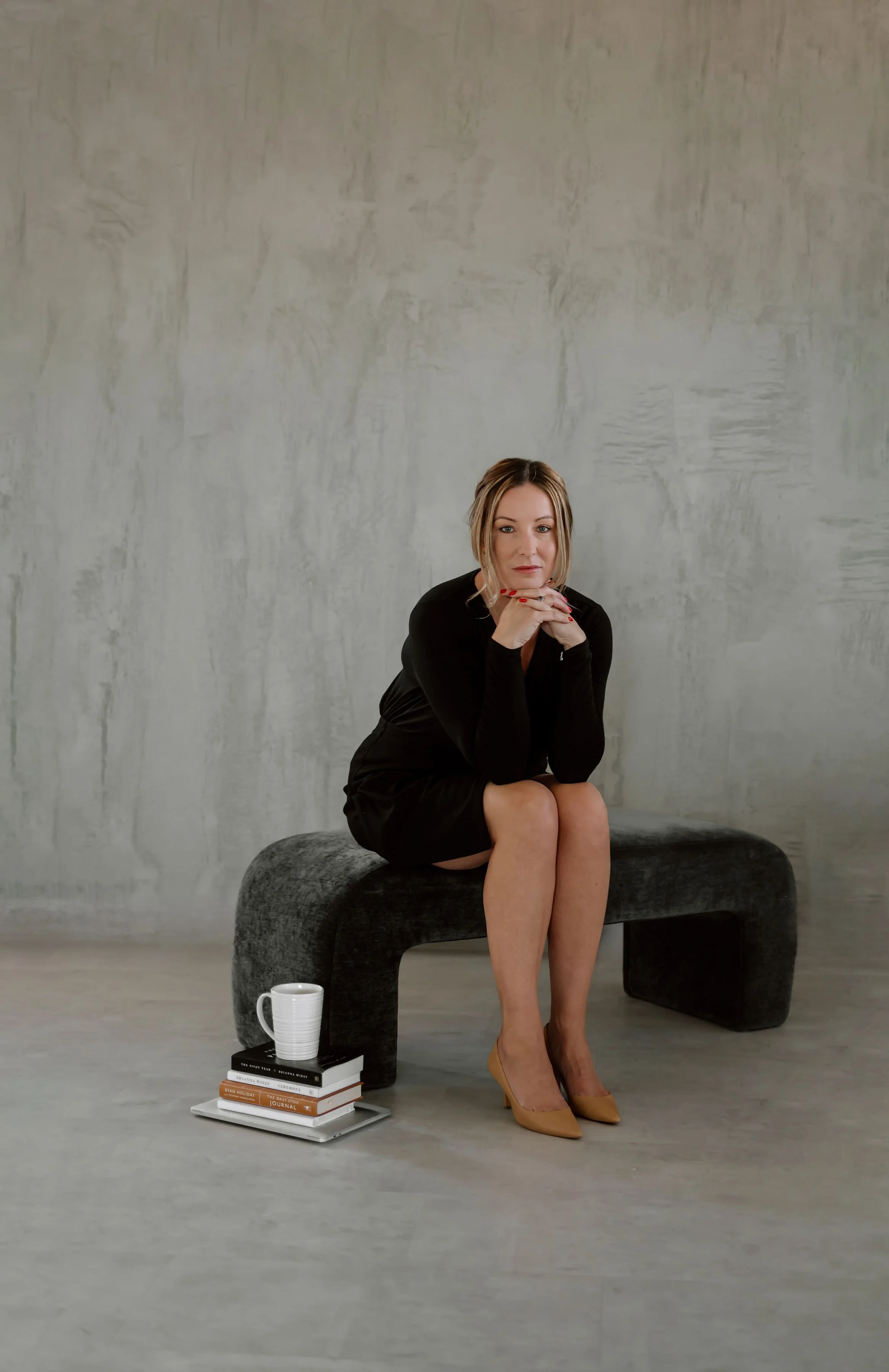 A portrait of Heather Foidart, sitting on a dark upholstered bench with her chin resting on her hands. She wears a black dress and beige high heels, with crossed legs. There is a cup on a stack of books and magazines on a tray in front of her.