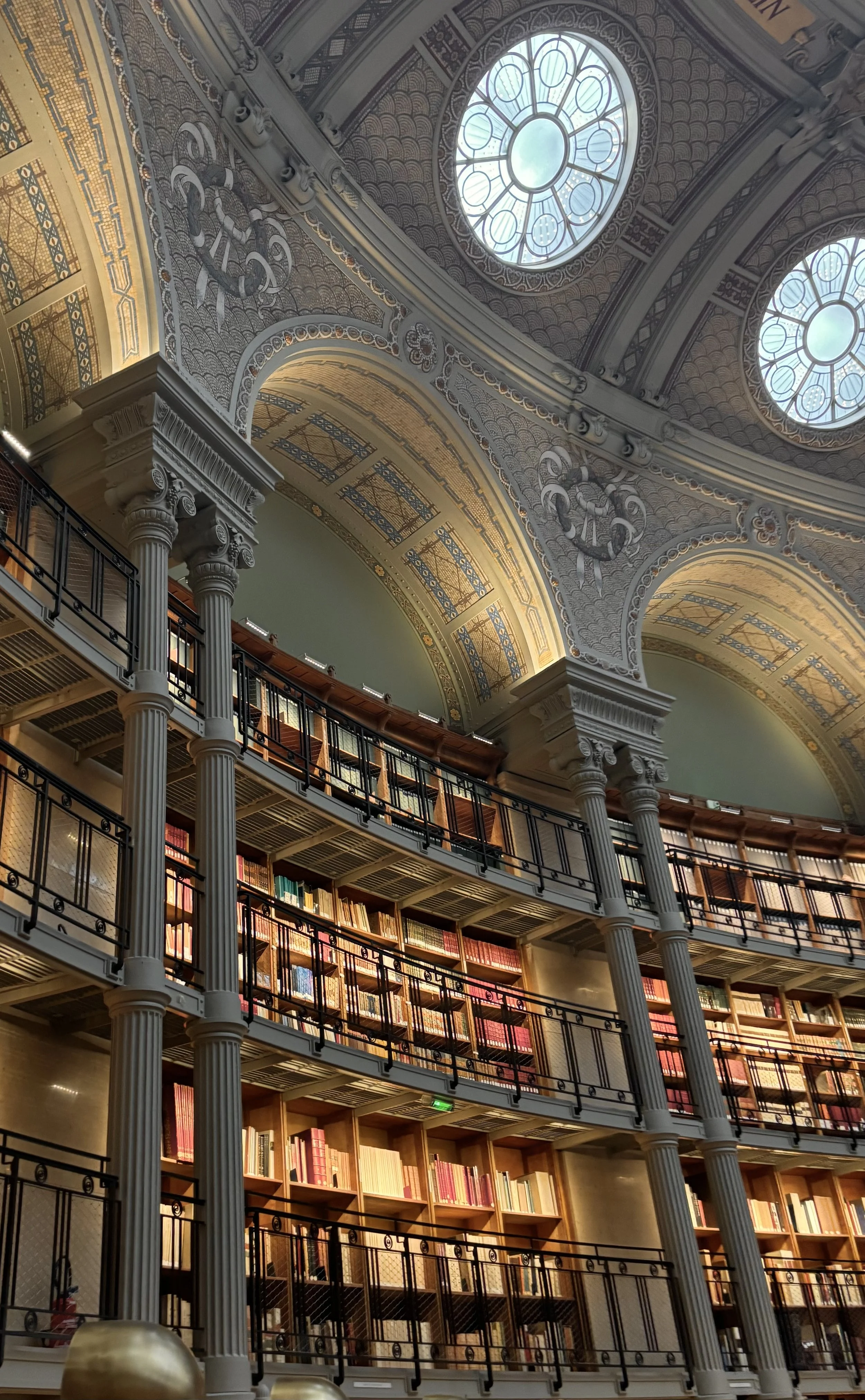 Interior view of a grand library with multiple levels of bookshelves, ornate columns, and large arched windows with stained glass on the ceiling.