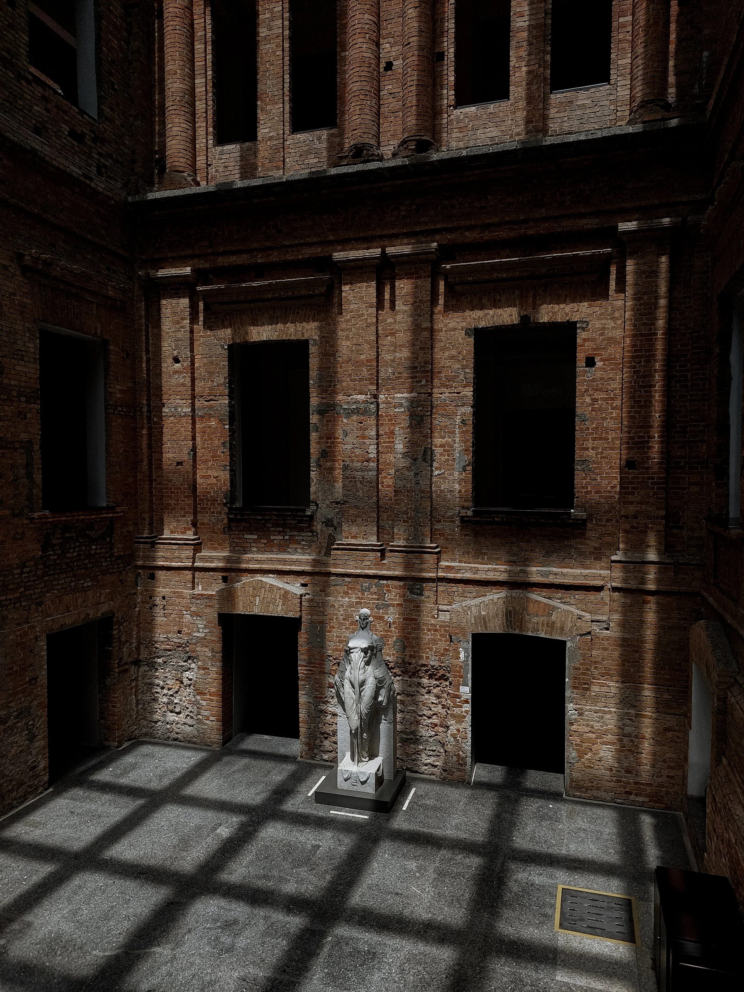 Interior of a brick building with multiple window openings, some open and some closed, a statue of a woman in the center, and casting shadows on the floor.