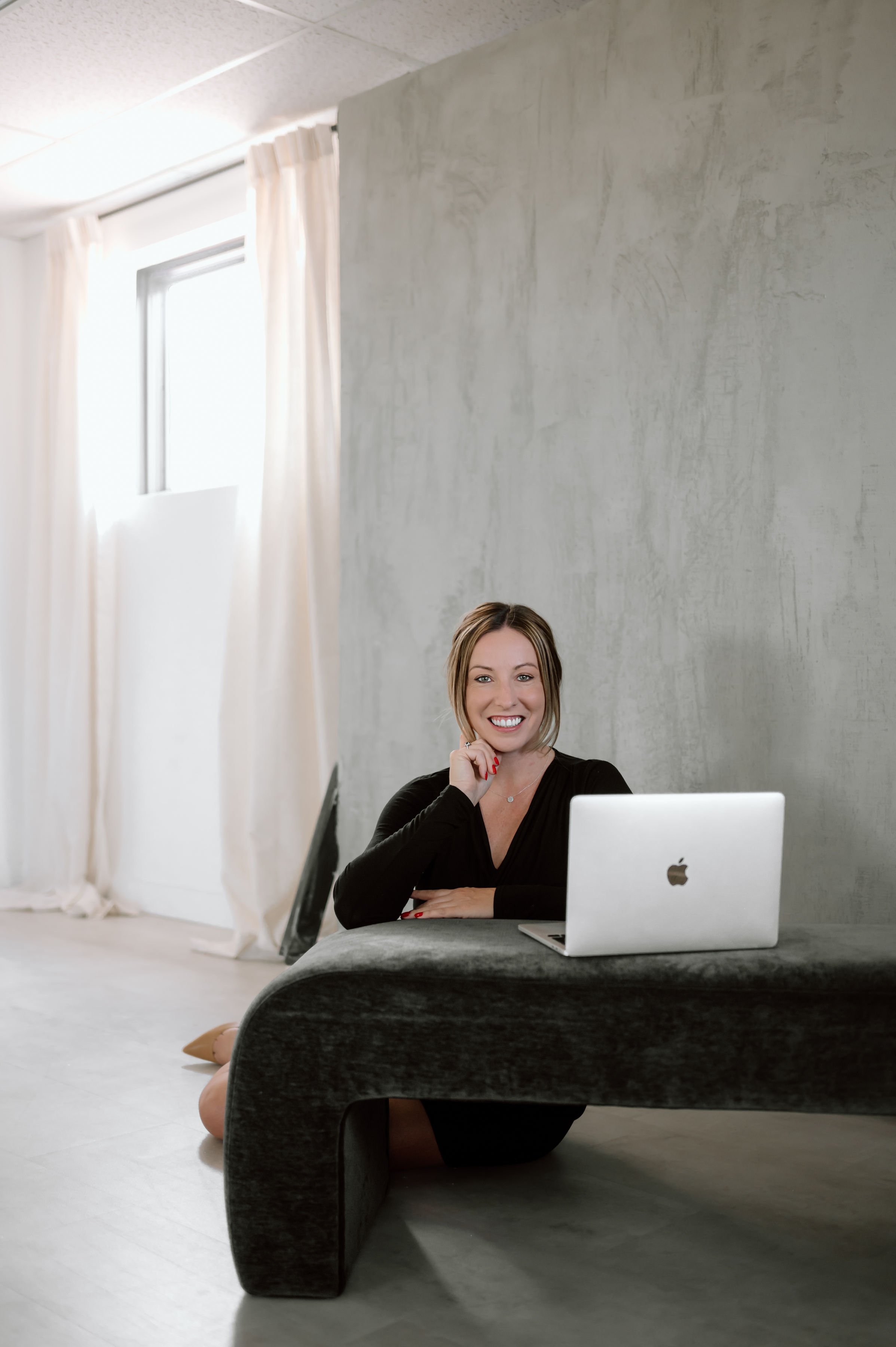 Heather Foidart, AZ, sitting on the floor behind a low black table with a gray tablecloth, smiling and looking at the camera, with an Apple laptop open in front of her. There is a computer monitor in front of the woman, partially visible.