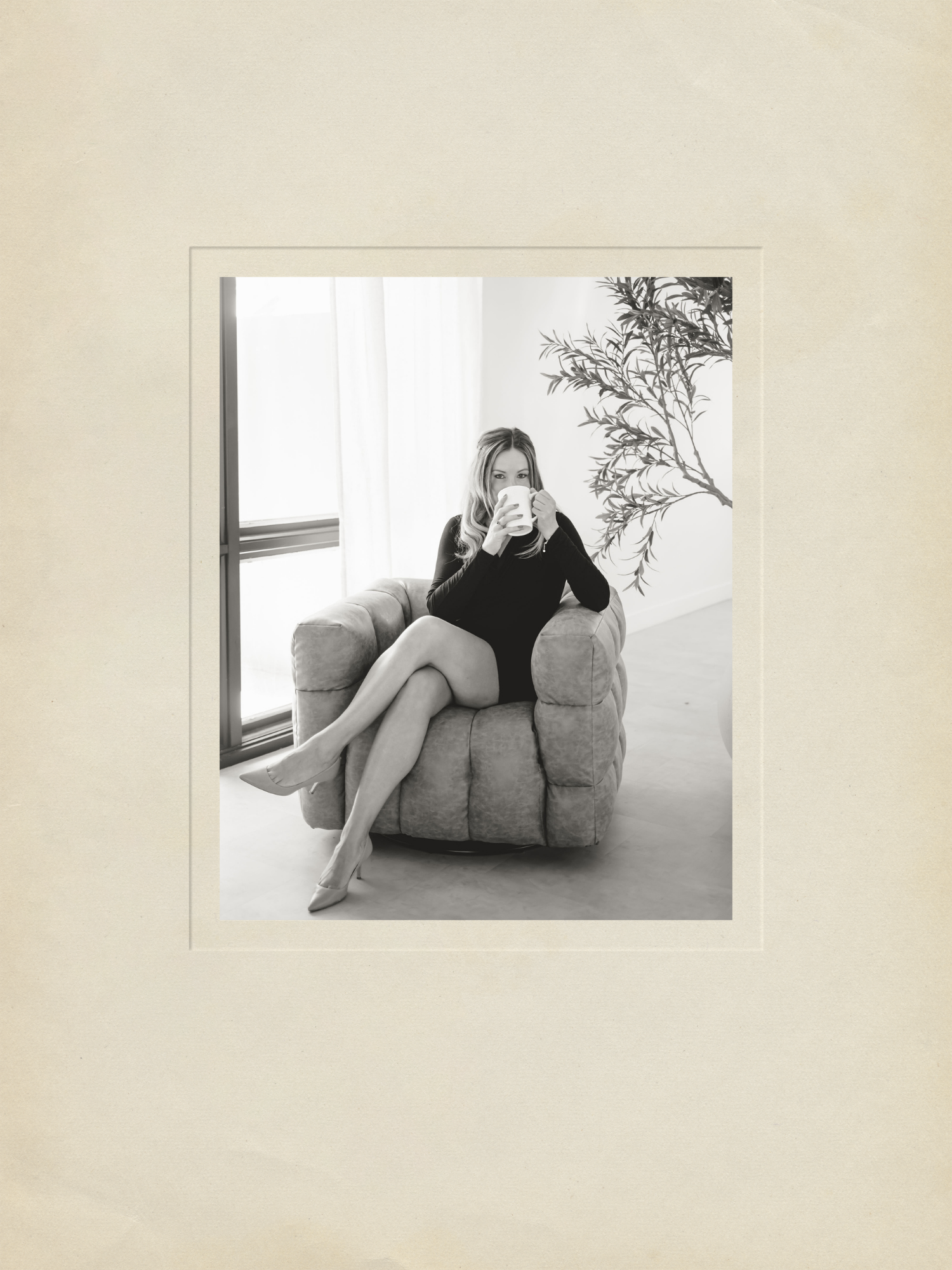 Heather Foidart, Corporate Advisor, sitting on a plush armchair, drinking from a mug in a bright room with large windows, a curtain, and a large plant.