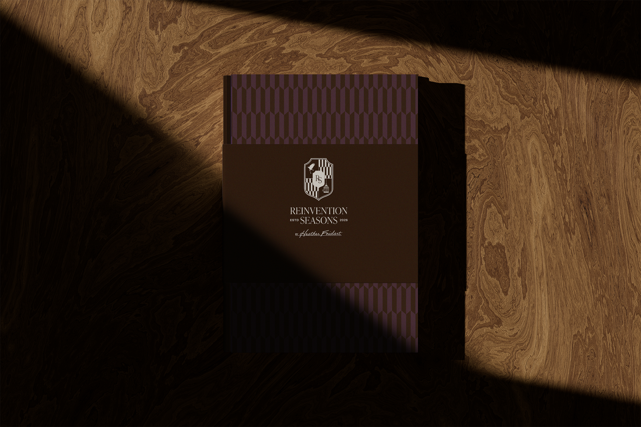 A n elegant book titled "REINVENTION SEASONS, the philosophy of seasonal intelligence" with geometric purple and brown pattern on the lid, placed on a wooden surface with shadows cast across it.