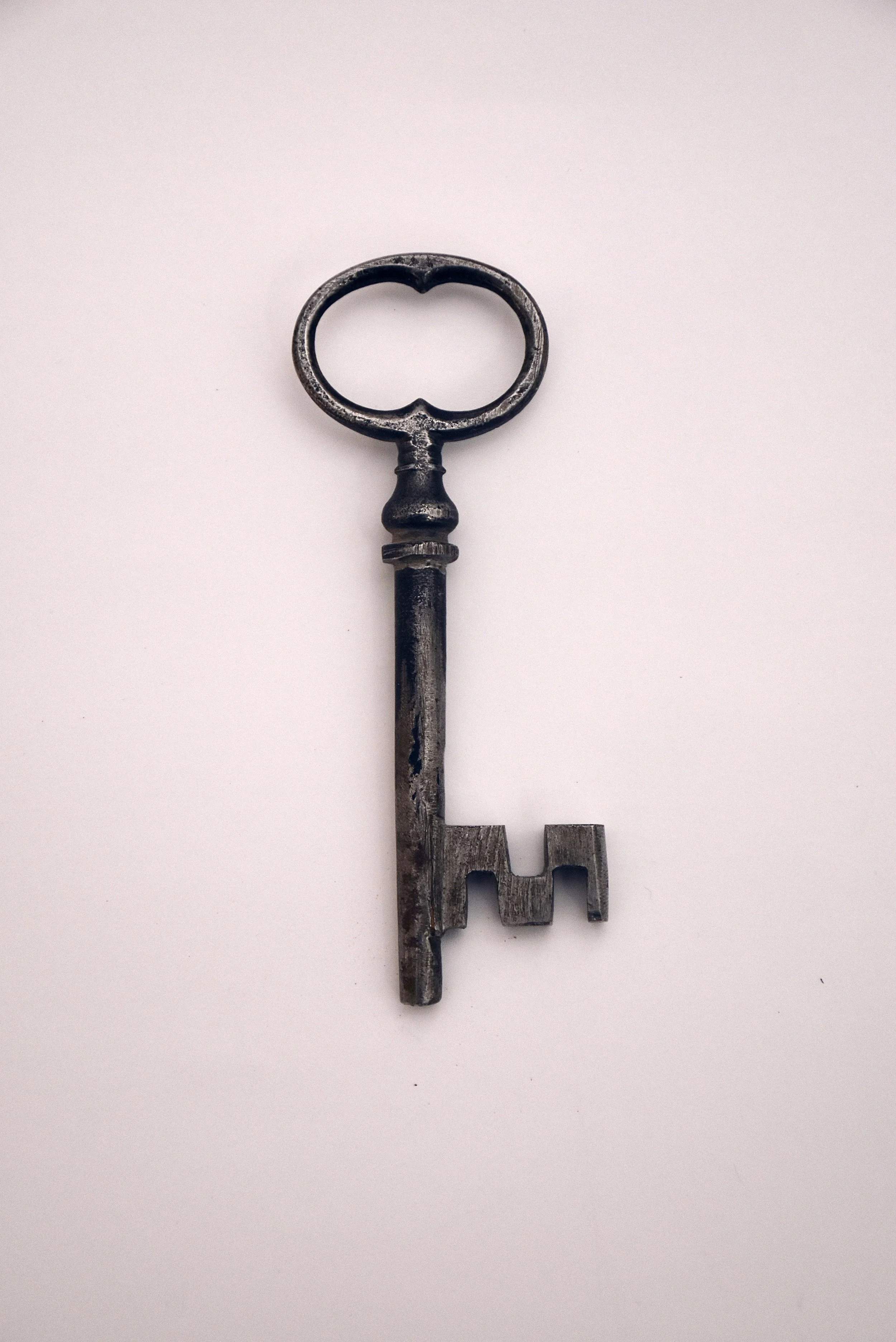 An antique metal key with a round handle, ornate design, and a long shaft, lying flat on a plain white background.