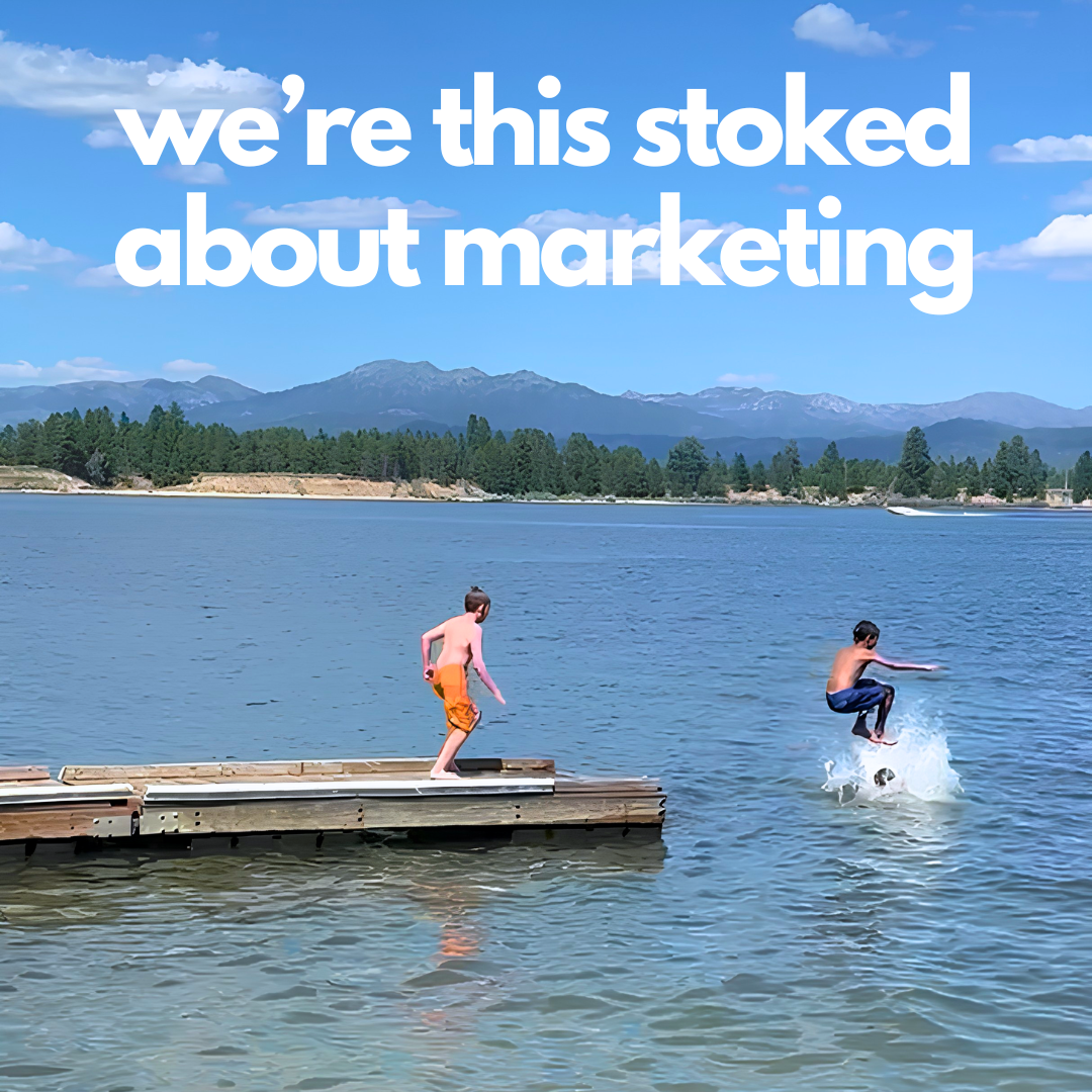 Two children playing near a lake with mountains in the background and a clear blue sky. One child is jumping into the water, and the other is standing on a wooden dock.