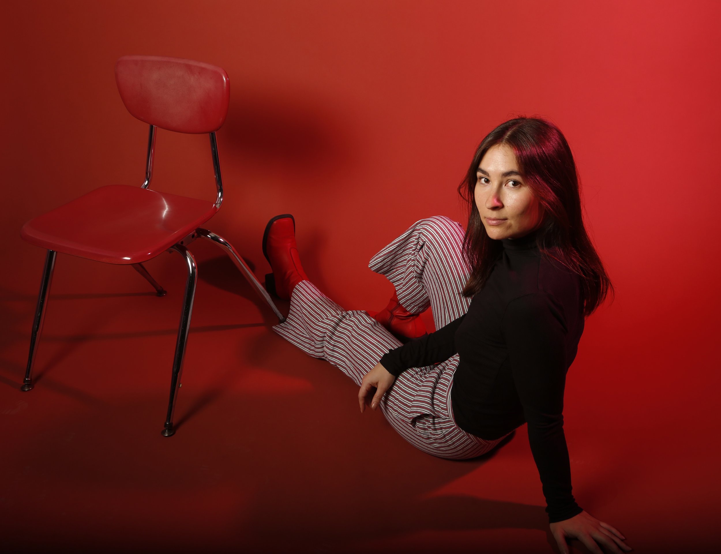 Image of Marie Vencil, an interior designer, sitting on a red floor against a red background, wearing striped pants and red boots, posing next to a red chair.
