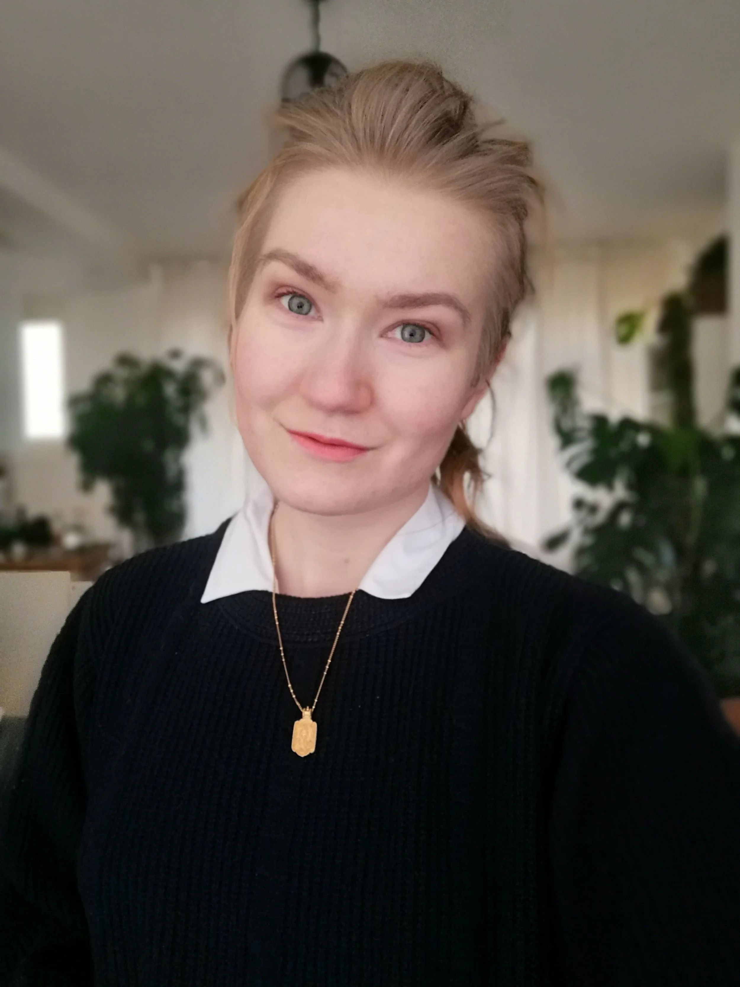 A young woman with blonde hair styled in an updo, wearing a white collared shirt under a black sweater, and a gold necklace with a pendant, smiling softly in an indoor setting with blurred plants and furniture in the background.