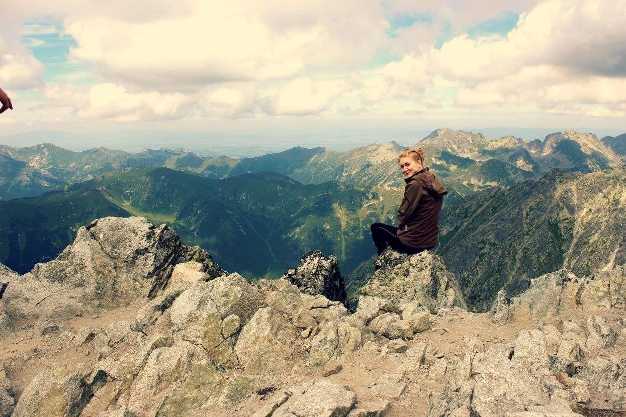 A woman sitting on a large rock on a mountain summit, overlooking green mountains with a cloudy sky above.