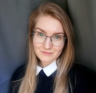Portrait of a young woman with long blonde hair, wearing glasses, a white shirt, and a dark sweater, standing against a gray background.