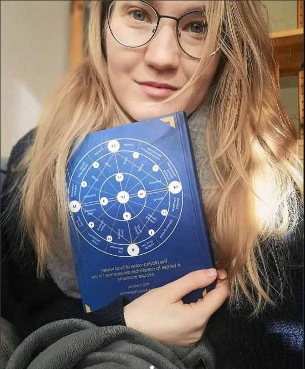 A young woman with long blonde hair, glasses, and a gray scarf, holding a blue circular chart or diagram close to her chest. She is smiling slightly and appears to be in a casual indoor setting.
