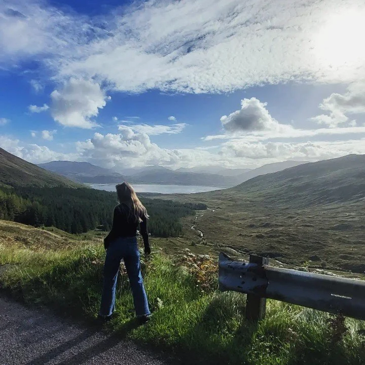 A woman with long hair, wearing a black top and blue jeans, stands on a roadside overlooking a scenic landscape. The view includes rolling hills, a river or lake, dense forests, and cloudy sky in the distance.