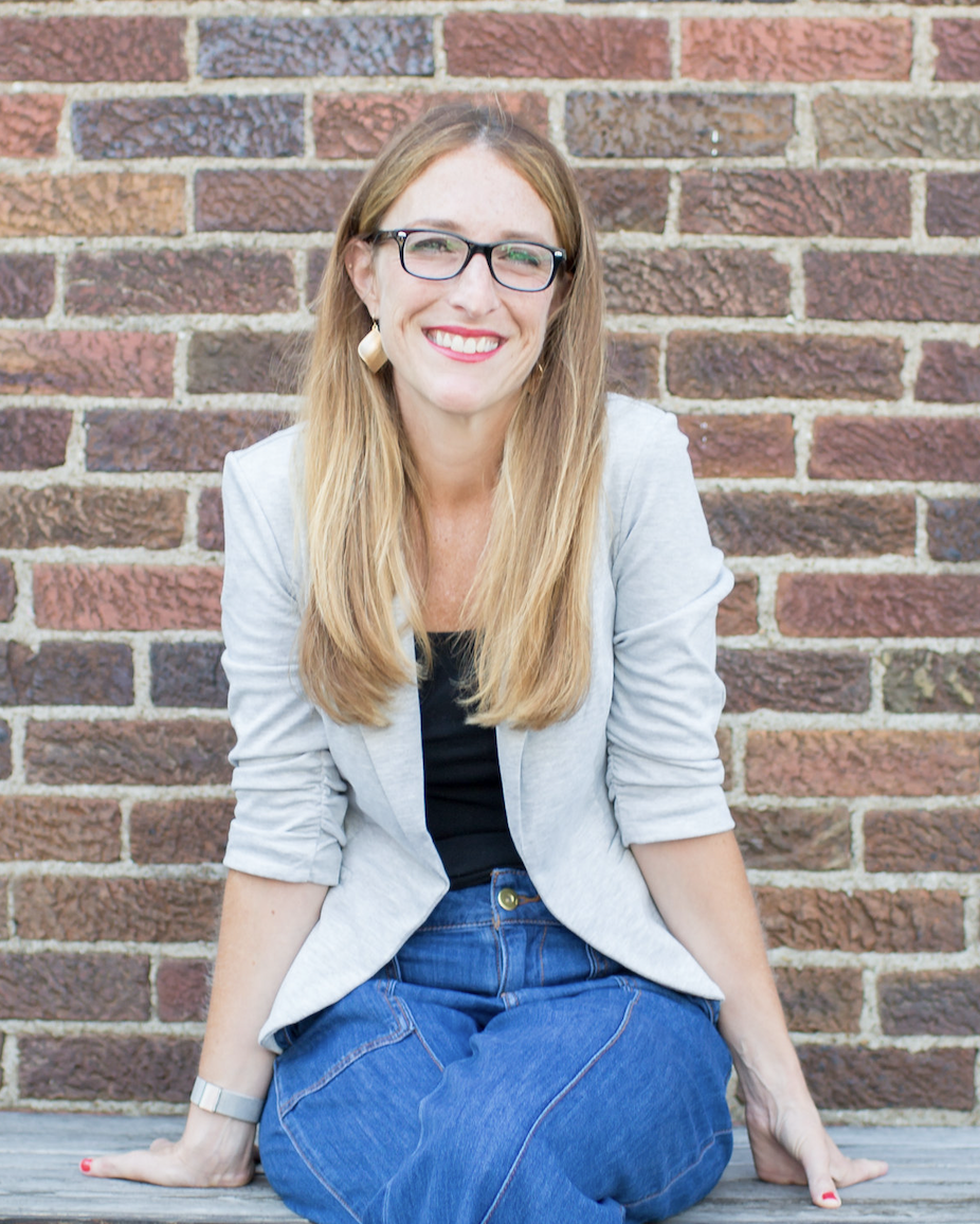 A woman with long blonde hair, glasses, and light skin sitting on the ground against a brick wall, smiling at the camera, wearing a light gray blazer, black shirt, and blue jeans.