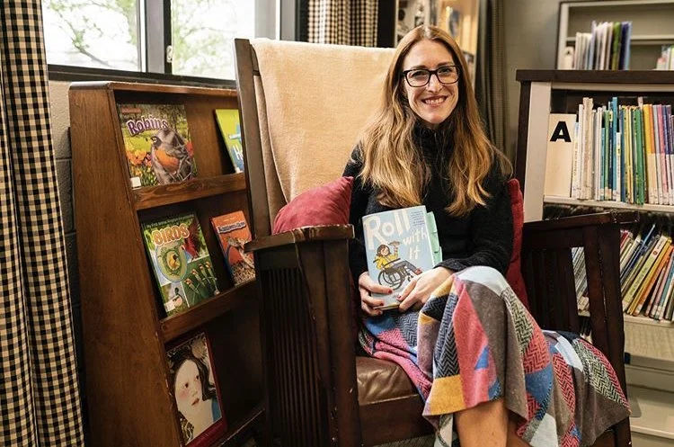A woman sitting in a cozy chair in a library, holding a book titled 'Pool With It'. She has long hair, glasses, and is smiling, with a multicolored blanket over her lap.