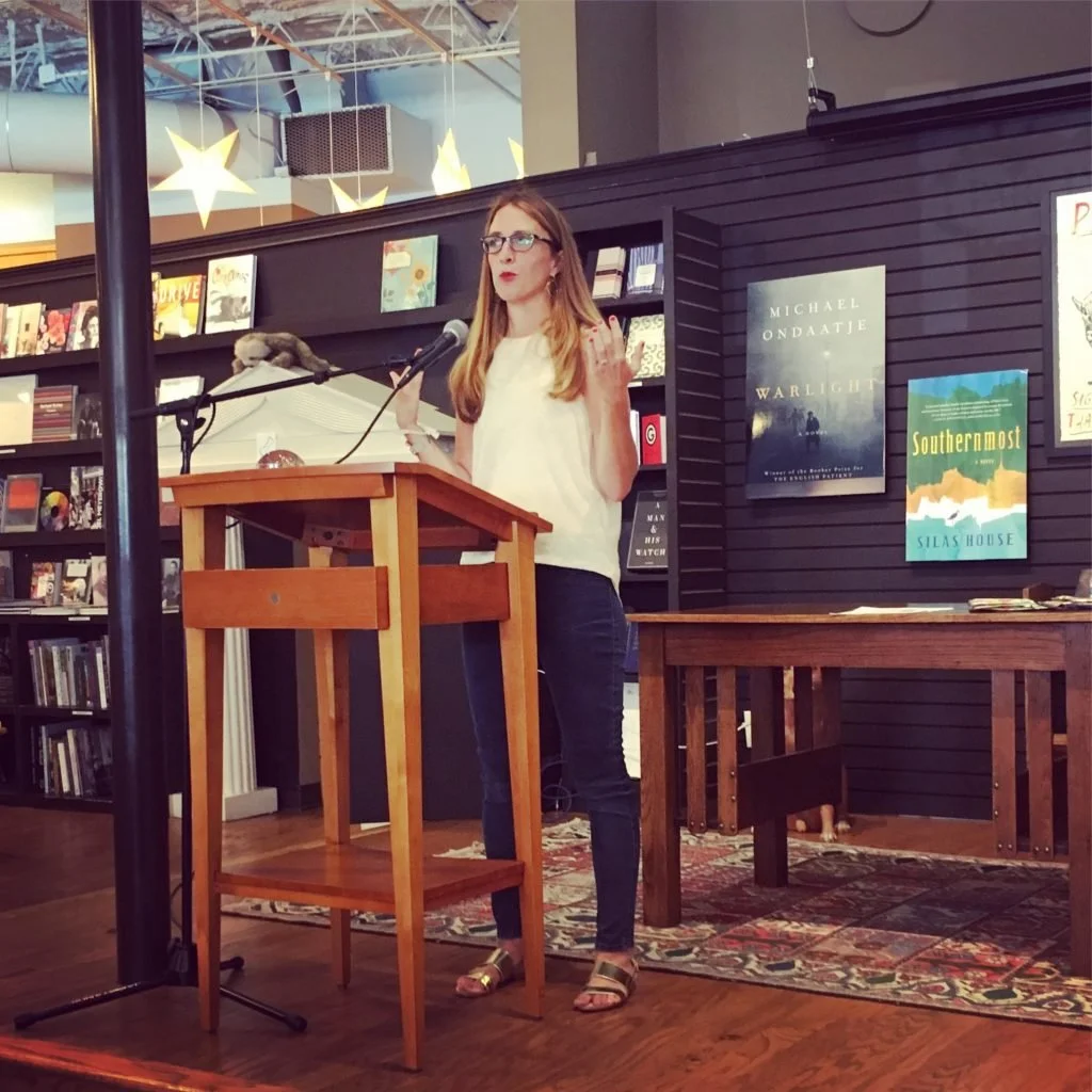 A woman with glasses and long strawberry hair speaks at a podium in a bookstore or library, with shelves of books behind her and posters on the wall.