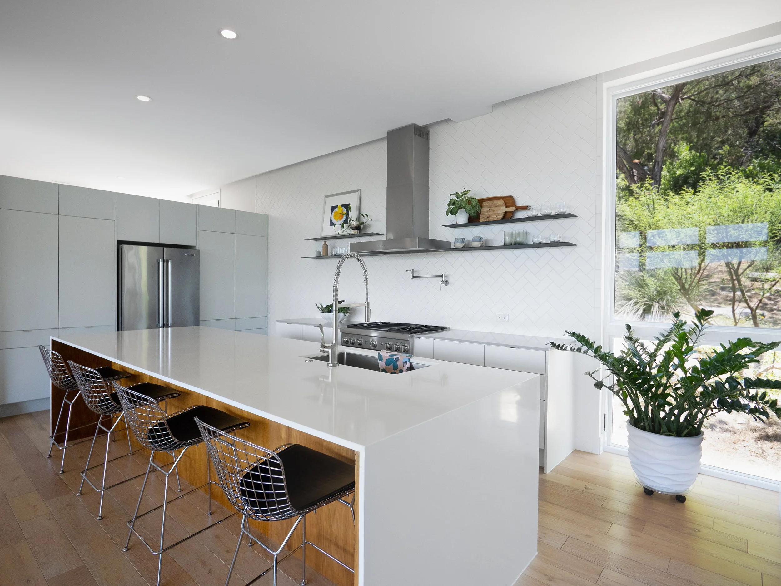 Modern kitchen with white island, stainless steel appliances, floating shelves with decor, large window showing greenery outside, and potted plant near the window.