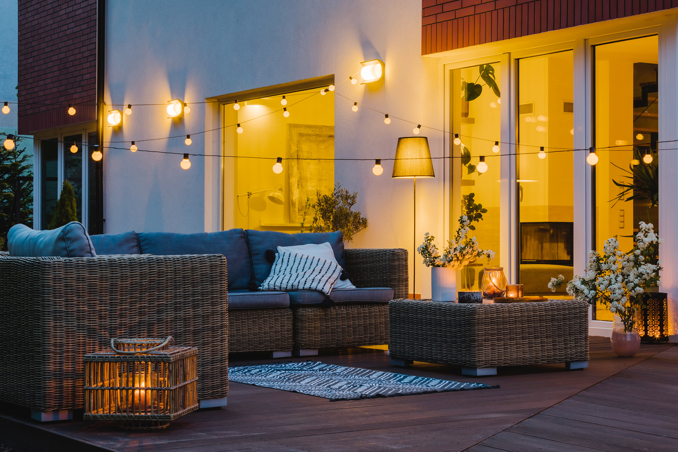 An outdoor patio at dusk with wicker furniture, a patterned outdoor rug, a tall floor lamp, potted plants, and string lights illuminating the area, adjacent to a house with large glass doors and interior lighting visible.