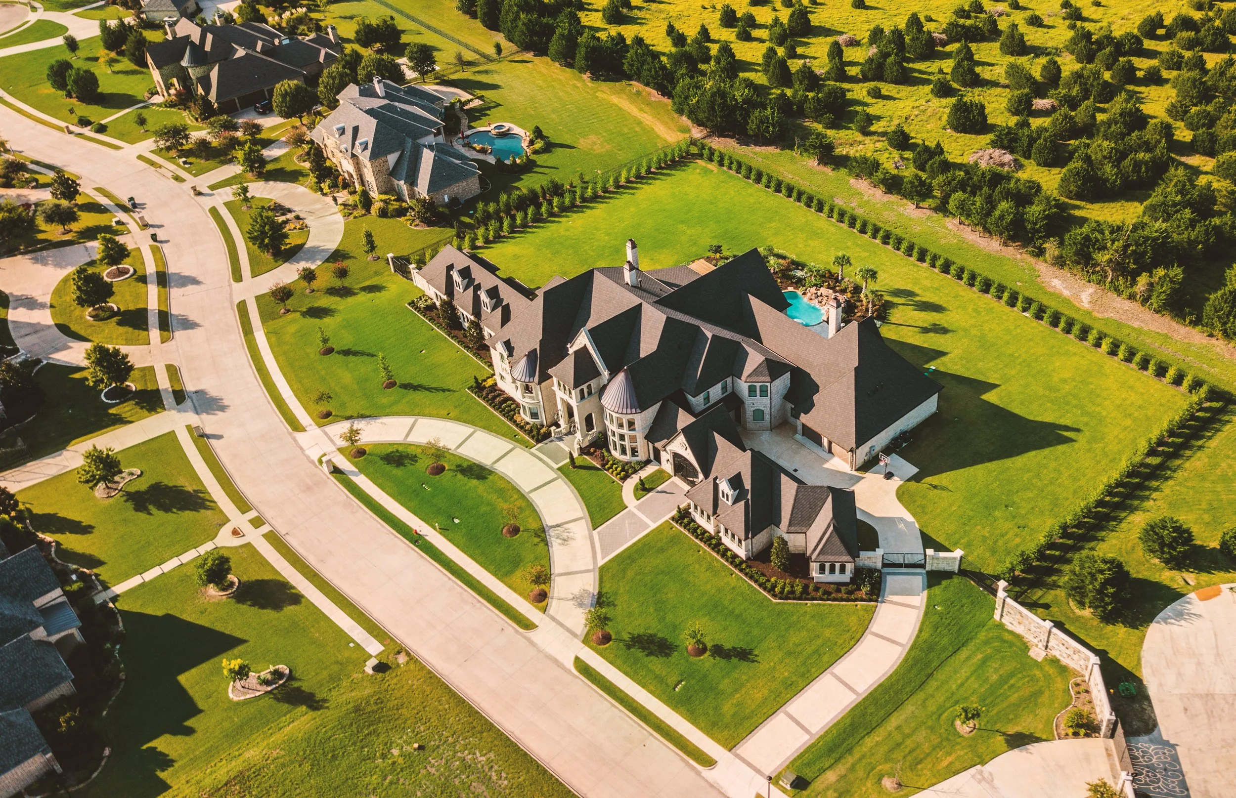 An aerial view of a suburban neighborhood showing multiple large houses with well-maintained lawns, driveways, and trees. Some houses have swimming pools in their backyards, and the area is surrounded by green open land and trees.