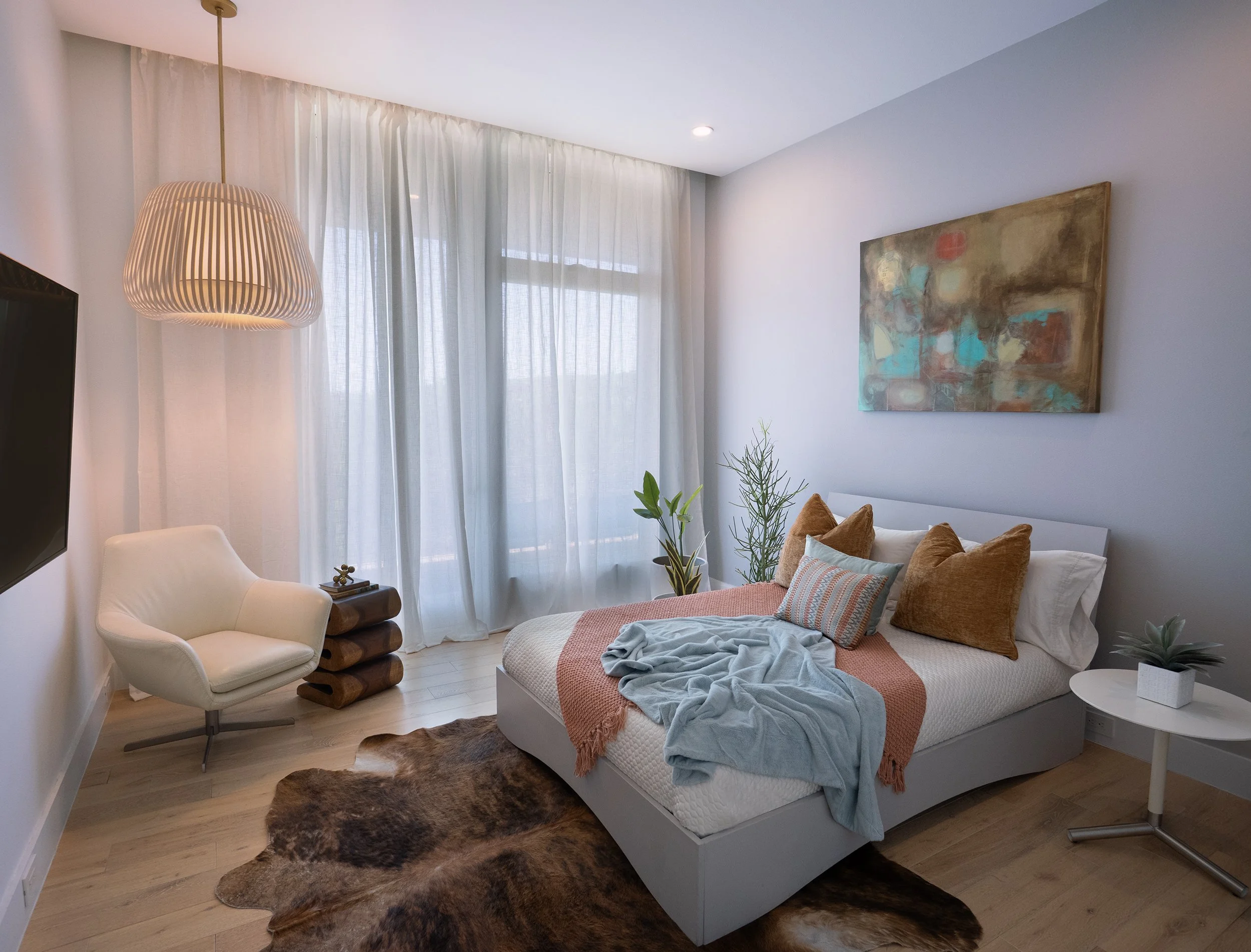 A bedroom with a bed, pillows, a blanket, and clothes draped over the bed. A cowhide rug is on the wooden floor. There is a white chair, a small table with books, houseplants, a large abstract painting on the wall, and a hanging light fixture near sheer curtains covering a window.