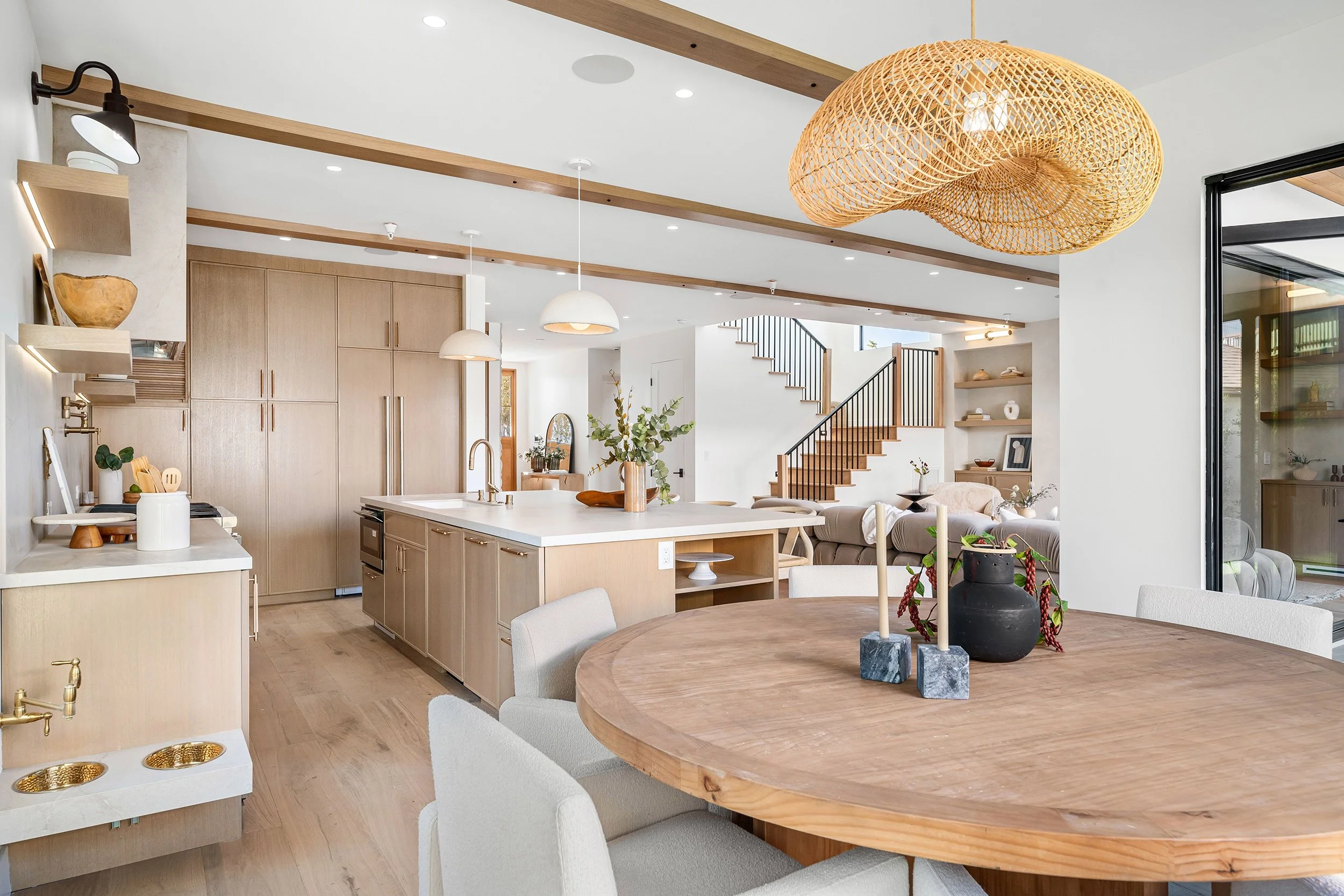 Open-concept kitchen and dining area with wooden accents, a large round wooden dining table, light-colored upholstered chairs, a kitchen island with a marble countertop, pendant and wall lighting, and a staircase with black railing in the background.